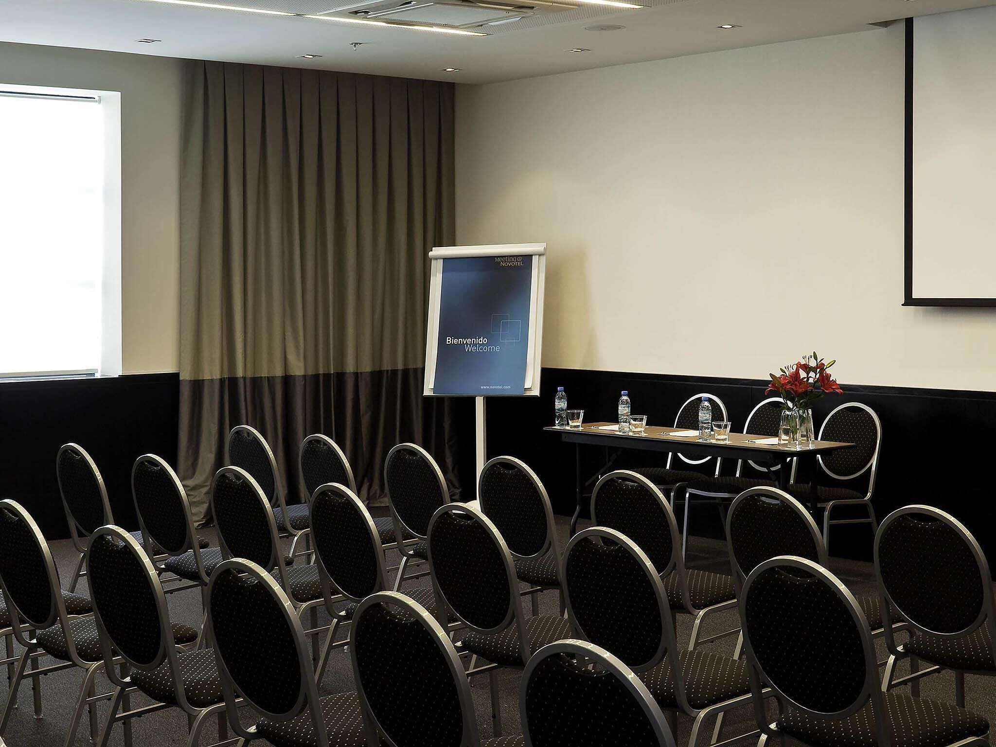 Ein Konferenzraum mit Reihen von Stühlen, einer Projektionsleinwand, einem Tisch mit Wasserflaschen und einem Vorhang.