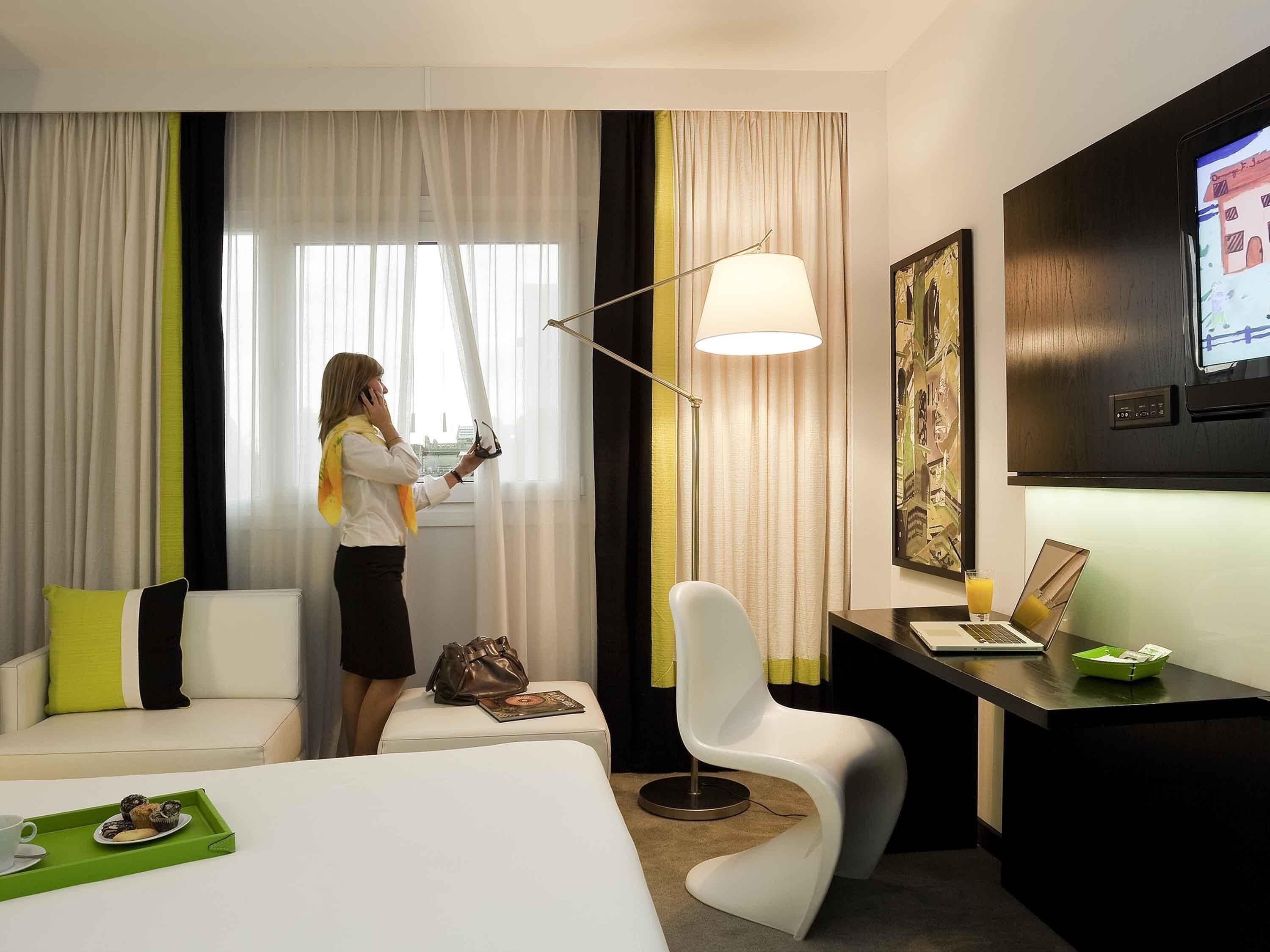 Quarto de hotel com cama, escrivaninha com laptop, cadeira, luminária e janela com cortinas.
