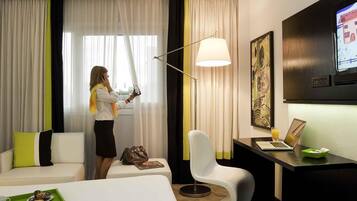 Quarto de hotel com cama, escrivaninha com laptop, cadeira, luminária e janela com cortinas.
