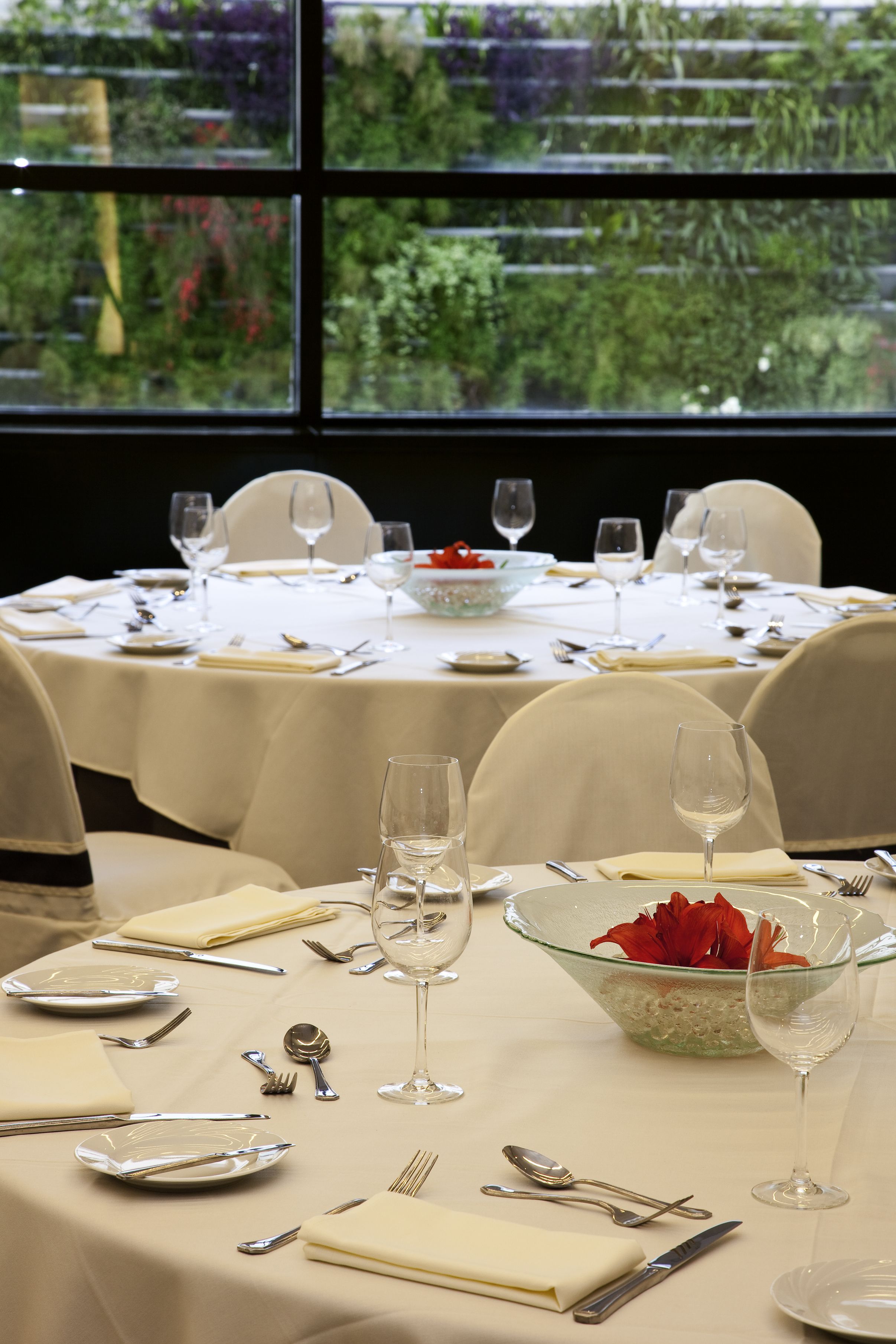 A dining table set for a meal with a bowl of red flowers, wine glasses, and cutlery.