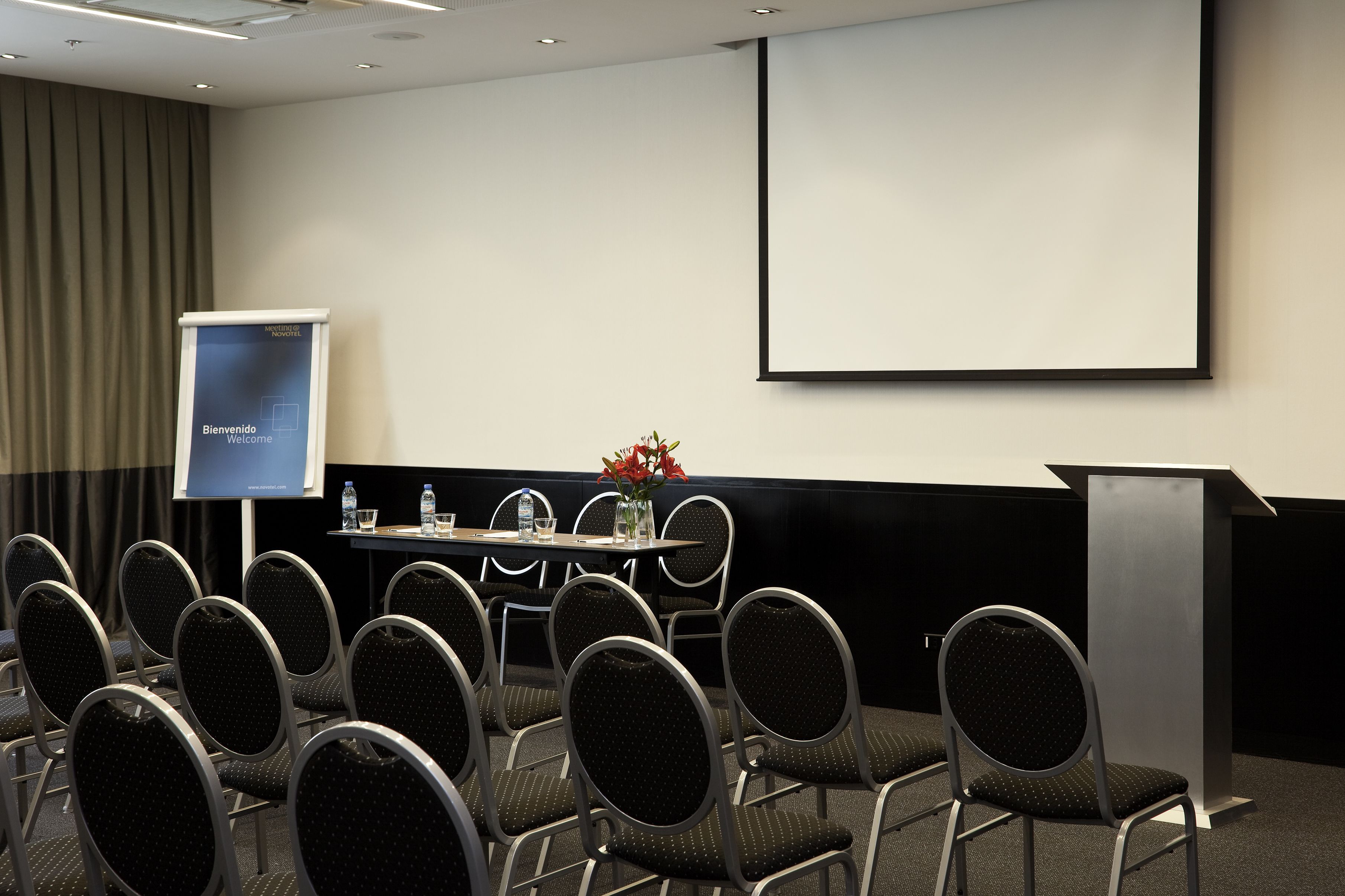 A conference room with rows of chairs, a podium, a screen, and a table with water bottles and glasses.