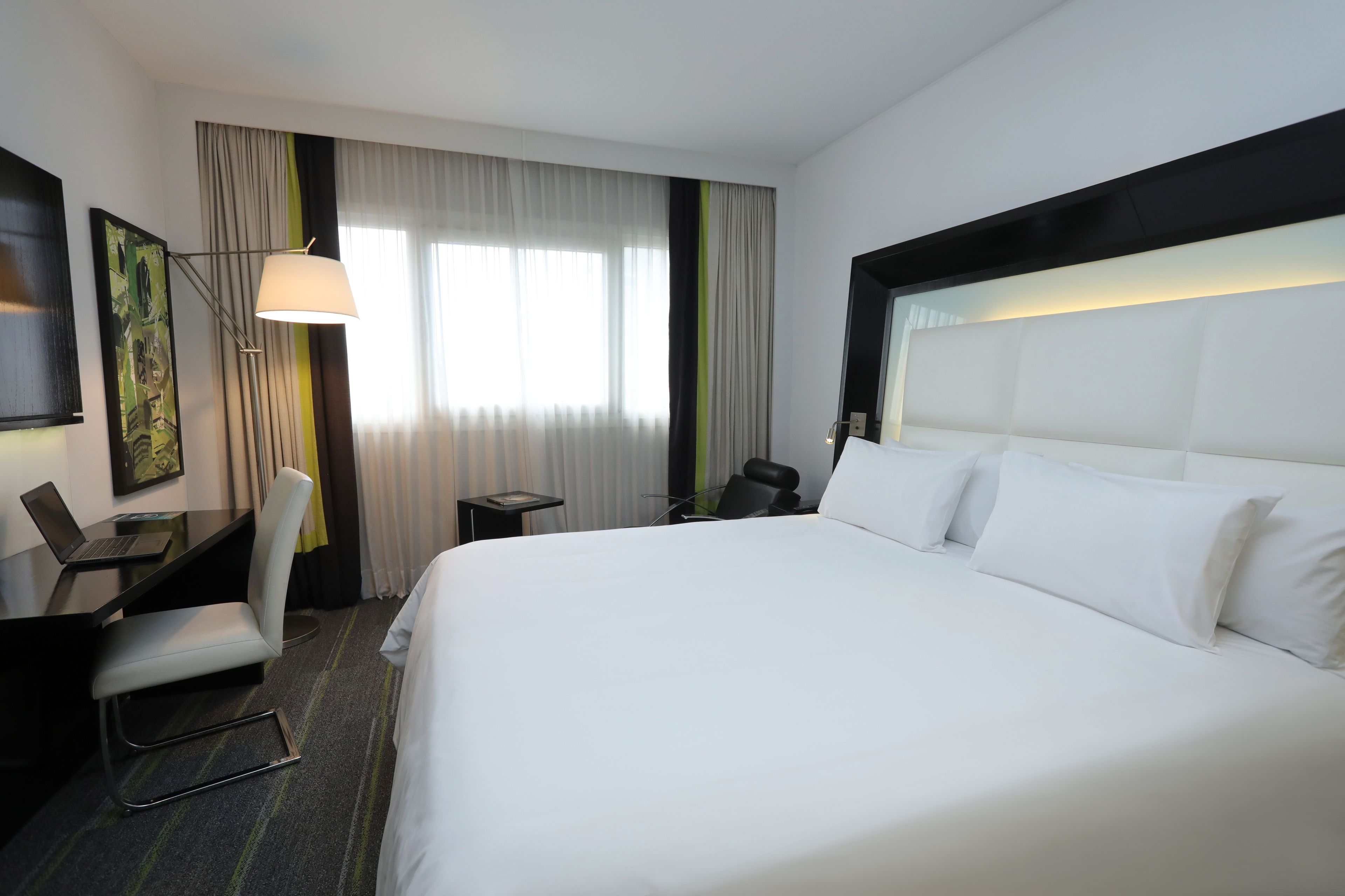 A hotel room with a large bed, a desk with a laptop, a chair, and a lamp.