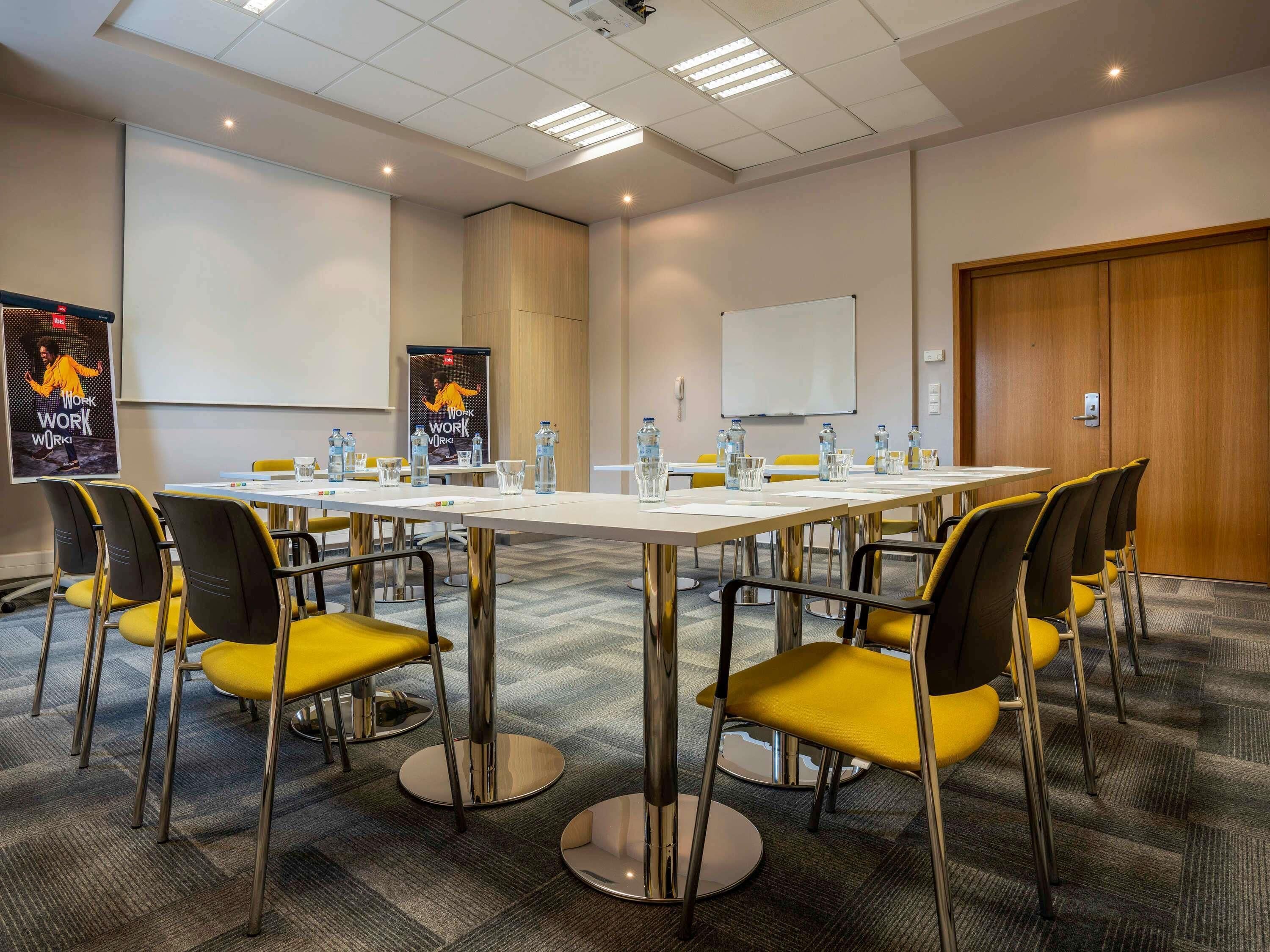 Une salle de conférence avec une longue table, des chaises jaunes, des bouteilles d’eau et un tableau blanc.