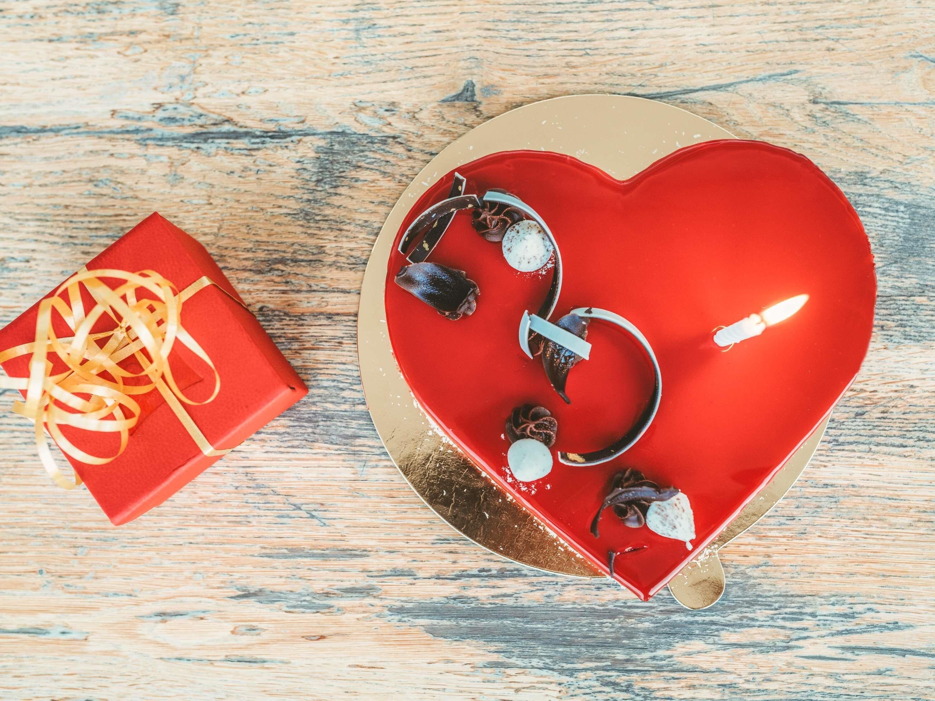 A red heart-shaped object with a gold rim on a wooden surface, a red gift box with a gold ribbon, and a small white object.