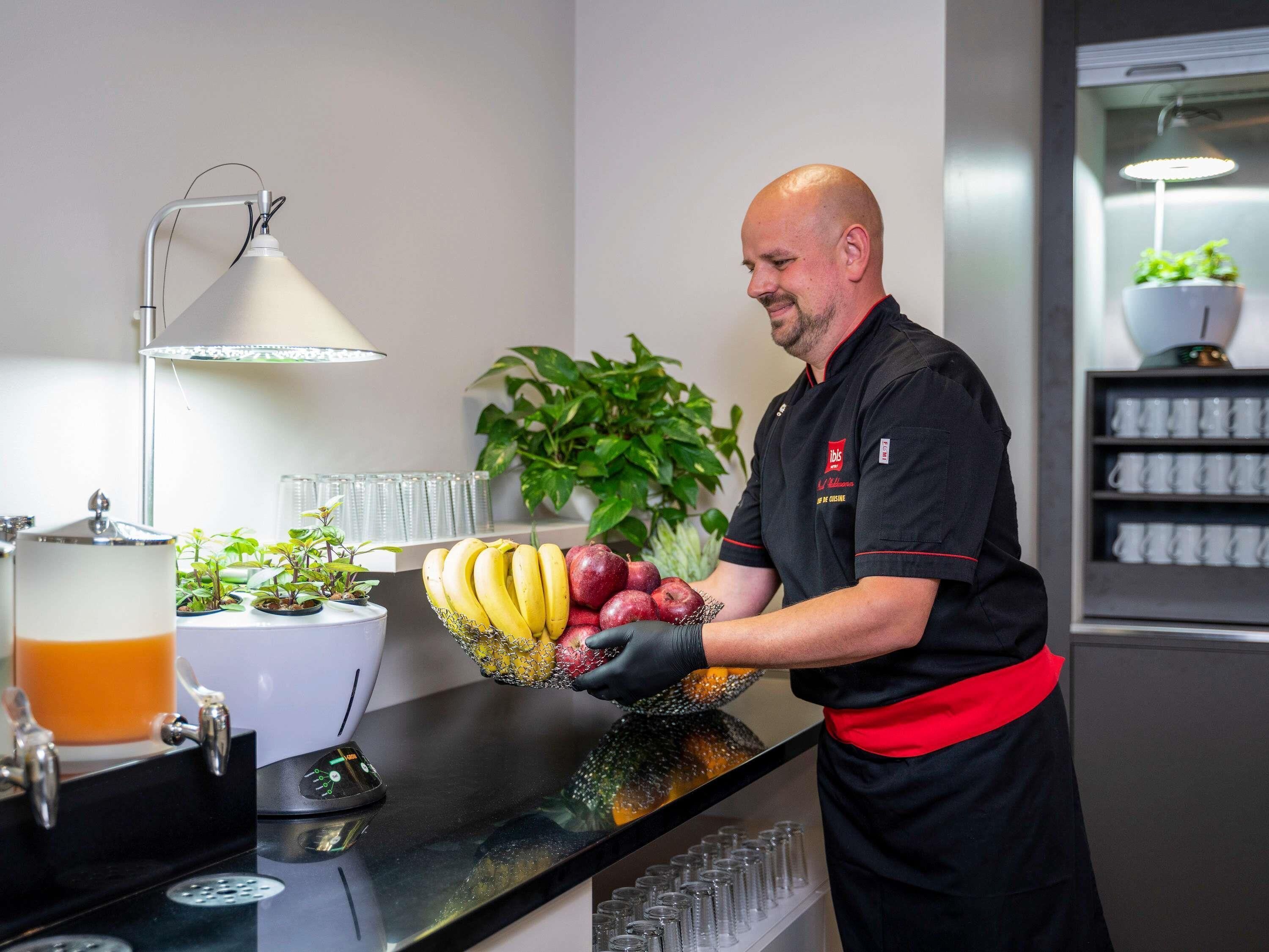 A person in a black uniform with a red apron, holding a bowl of fruit and bananas, standing in a kitchen area with a coffee machine, a plant, and a shelf with cups.