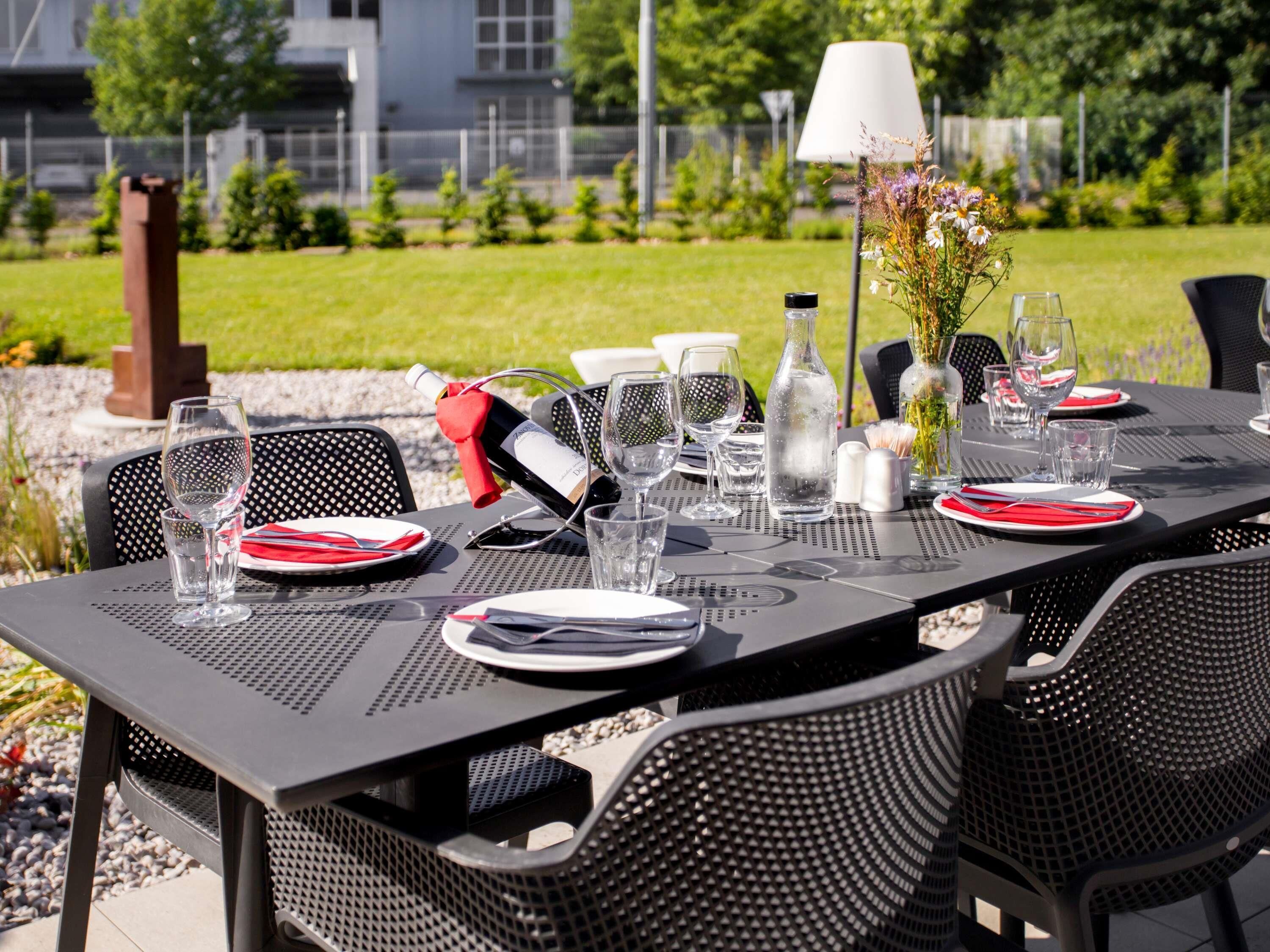 An outdoor dining area with tables set for two, featuring metal chairs, clear glassware, and a bottle of wine.