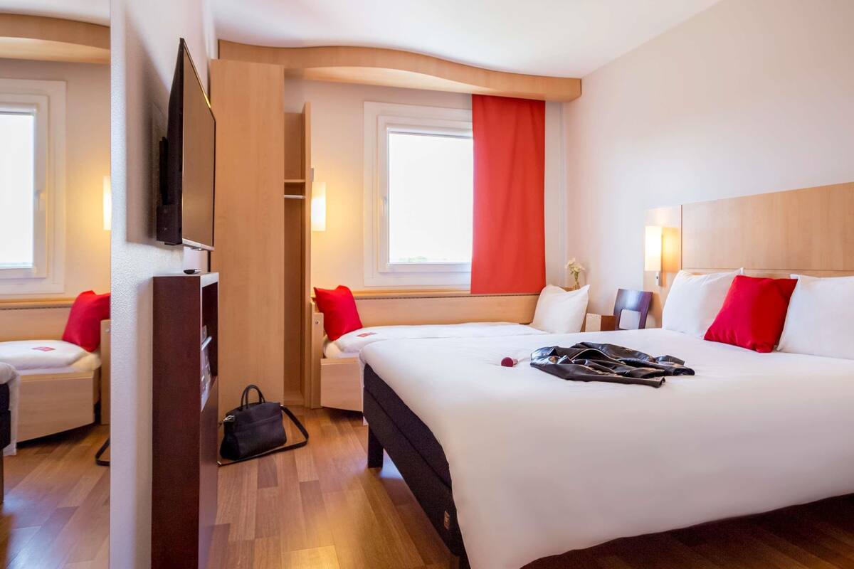 Ein Hotelzimmer mit einem großen Bett, einem Fernseher, einem Kleiderschrank und einem Fenster mit roten Vorhängen.