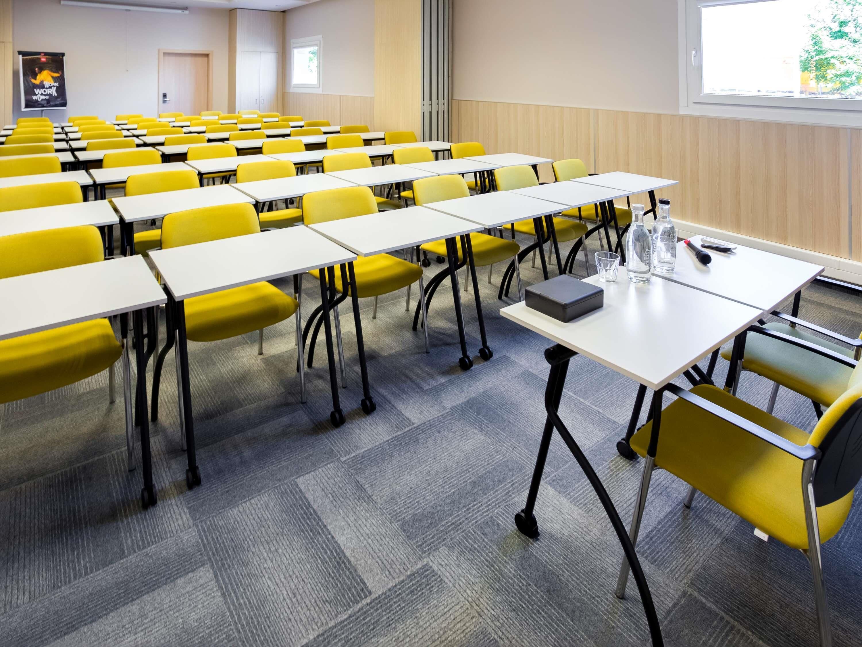 A conference room with rows of yellow chairs and white tables, carpeted floor, and a projector screen in the background.