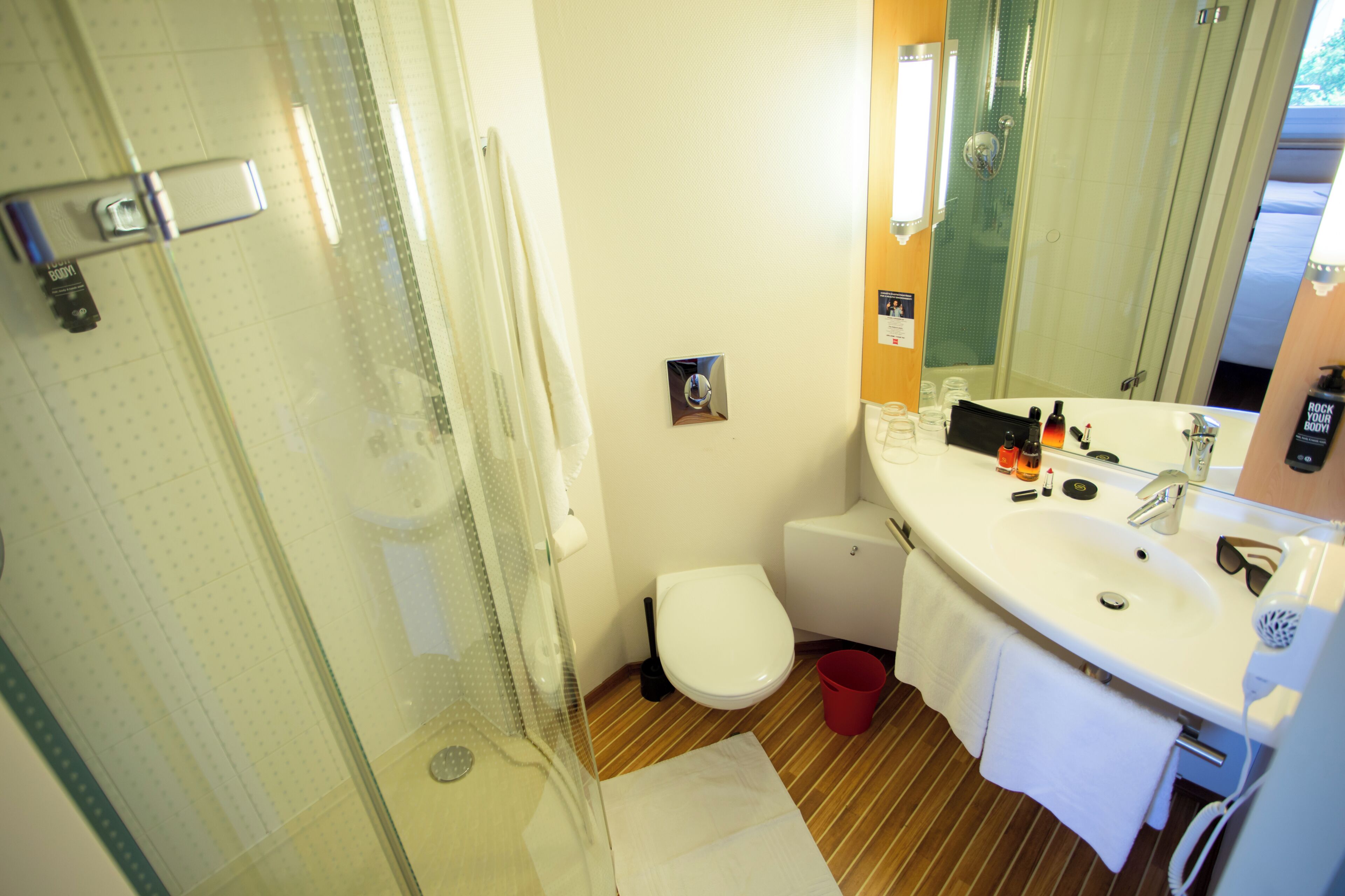 A bathroom with a glass shower enclosure, a white toilet, a sink with a mirror, and a red bucket.