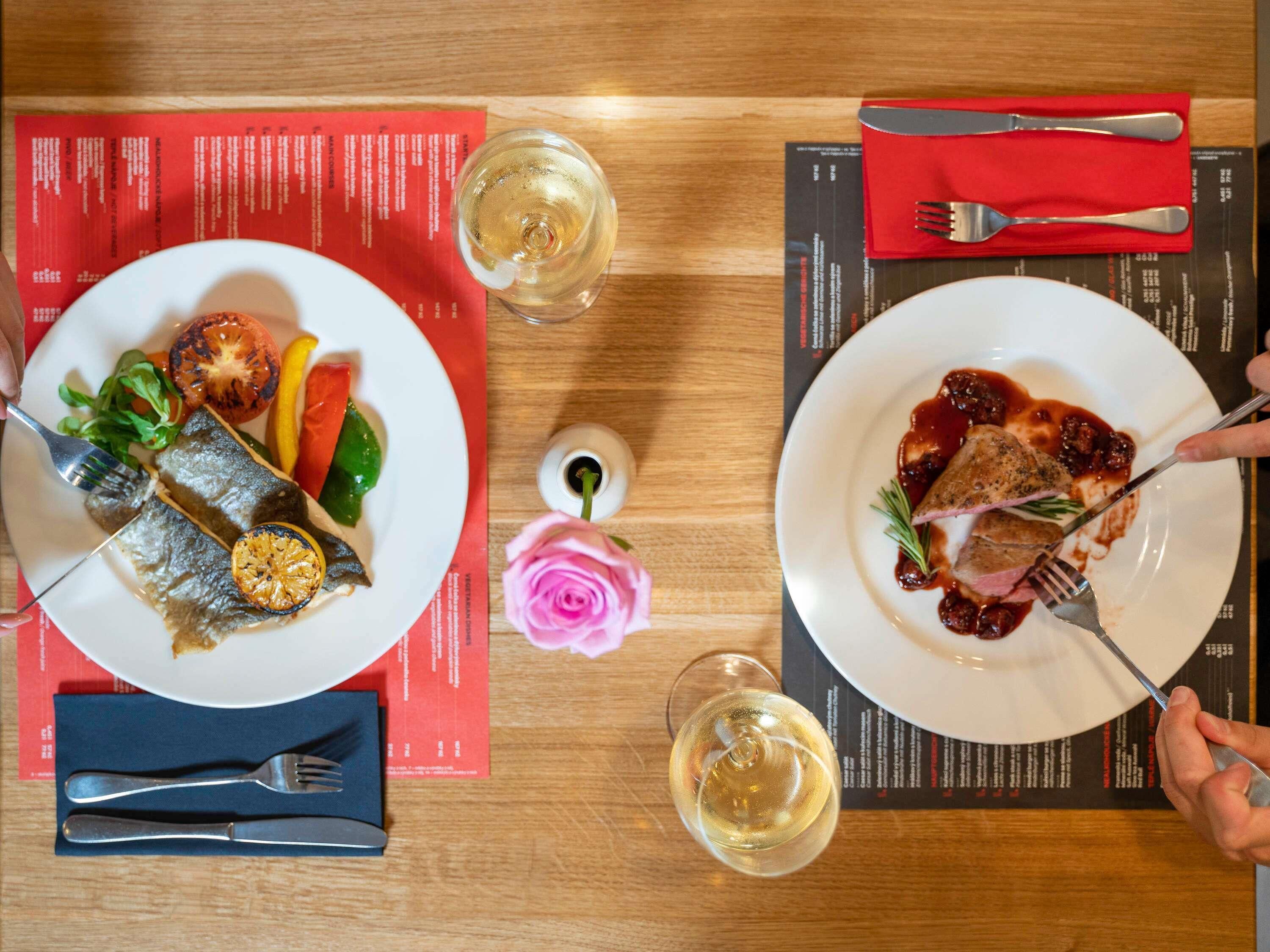 Two plates of food, a fork, a knife, and a glass of white wine on a wooden table.