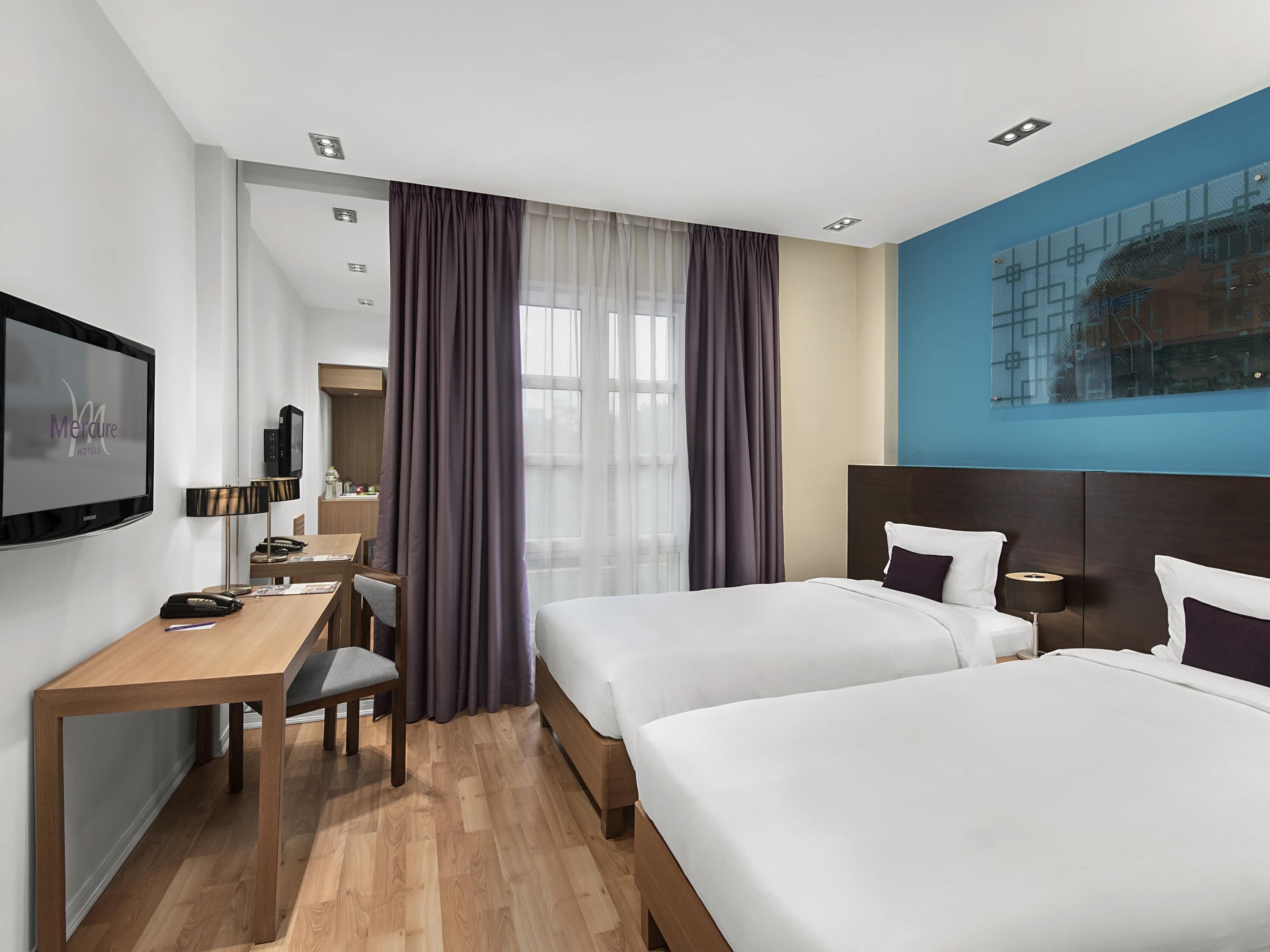 Hotellihuone, jossa on kaksi sänkyä, työpöytä, televisio ja suuri ikkuna, jossa on verhot.