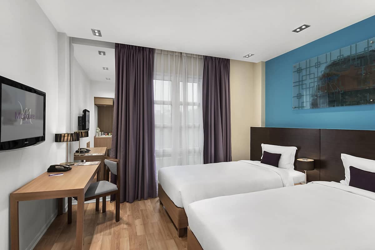 Hotellihuone, jossa on kaksi sänkyä, työpöytä, televisio ja suuri ikkuna, jossa on verhot.