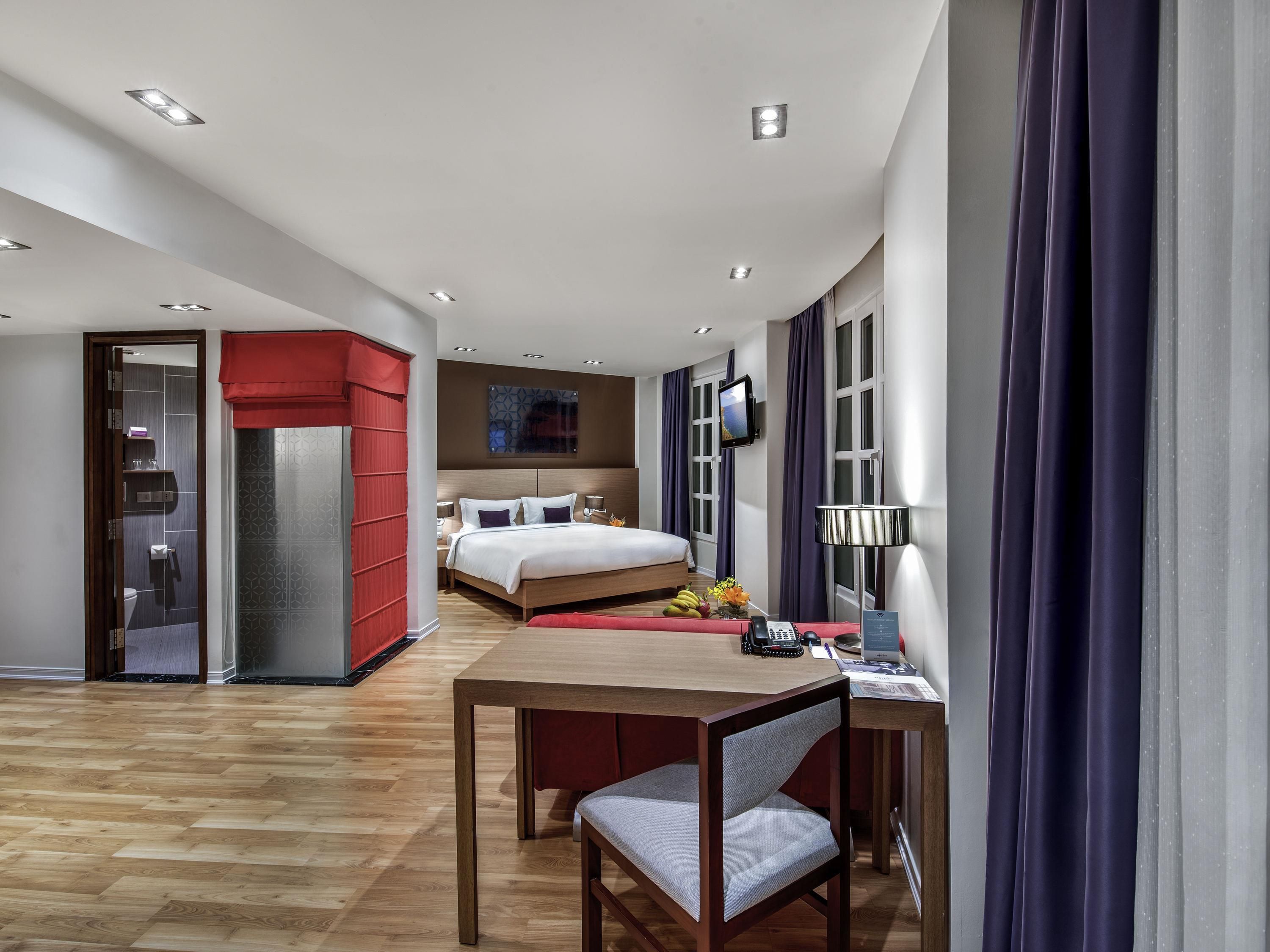 Una habitación de hotel moderna con suelo de madera, una cama con cabecero rojo, un escritorio con silla y un televisor montado en la pared.