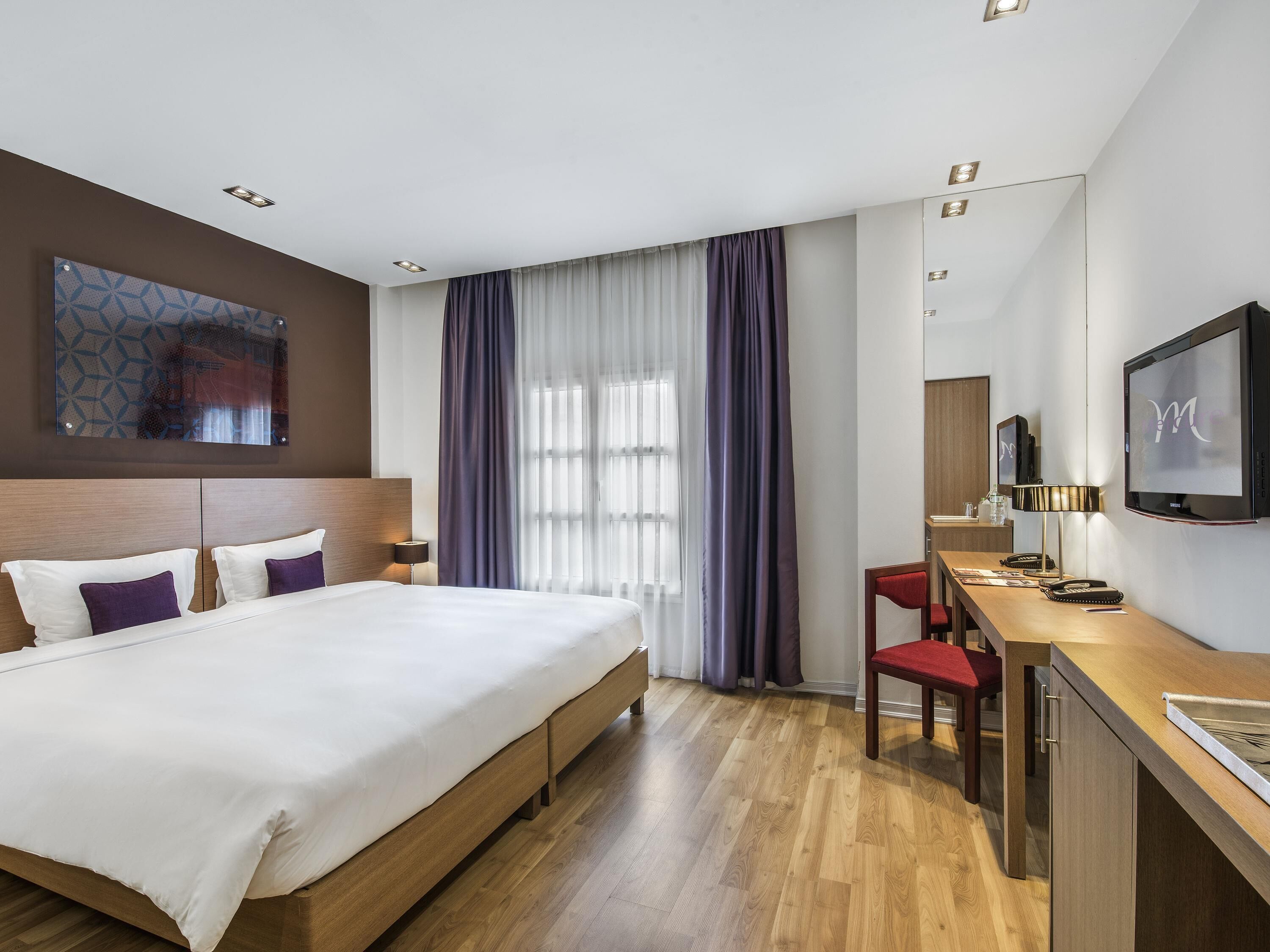 Hotellihuone, jossa on sänky, työpöytä, tuoli, televisio ja ikkuna, jossa on verhot.