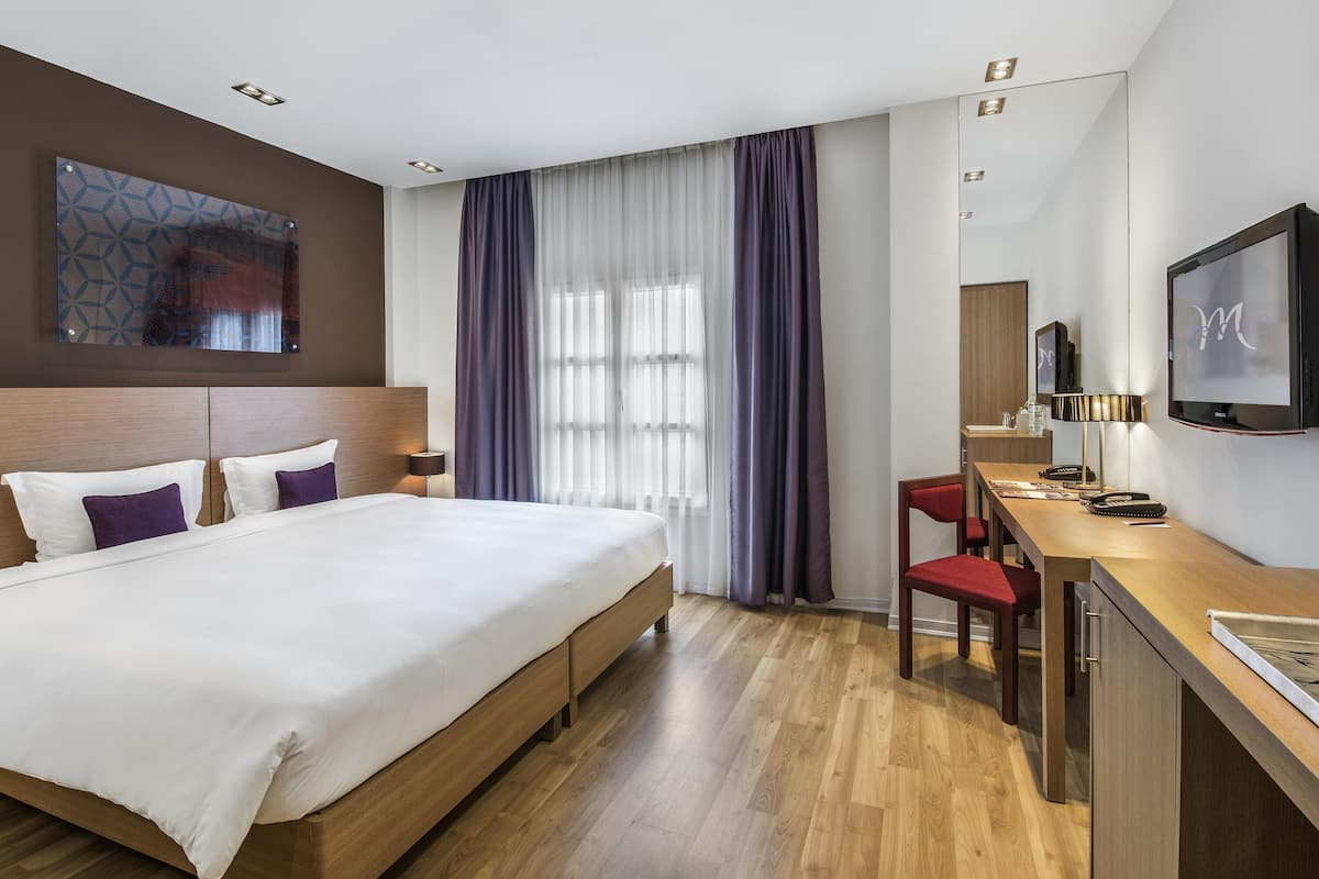 Hotellihuone, jossa on sänky, työpöytä, tuoli, televisio ja ikkuna, jossa on verhot.