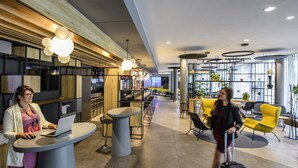 A modern hotel lobby with round tables, a bar area, and a seating area with yellow chairs. - ibis Muenchen City West (Munich)