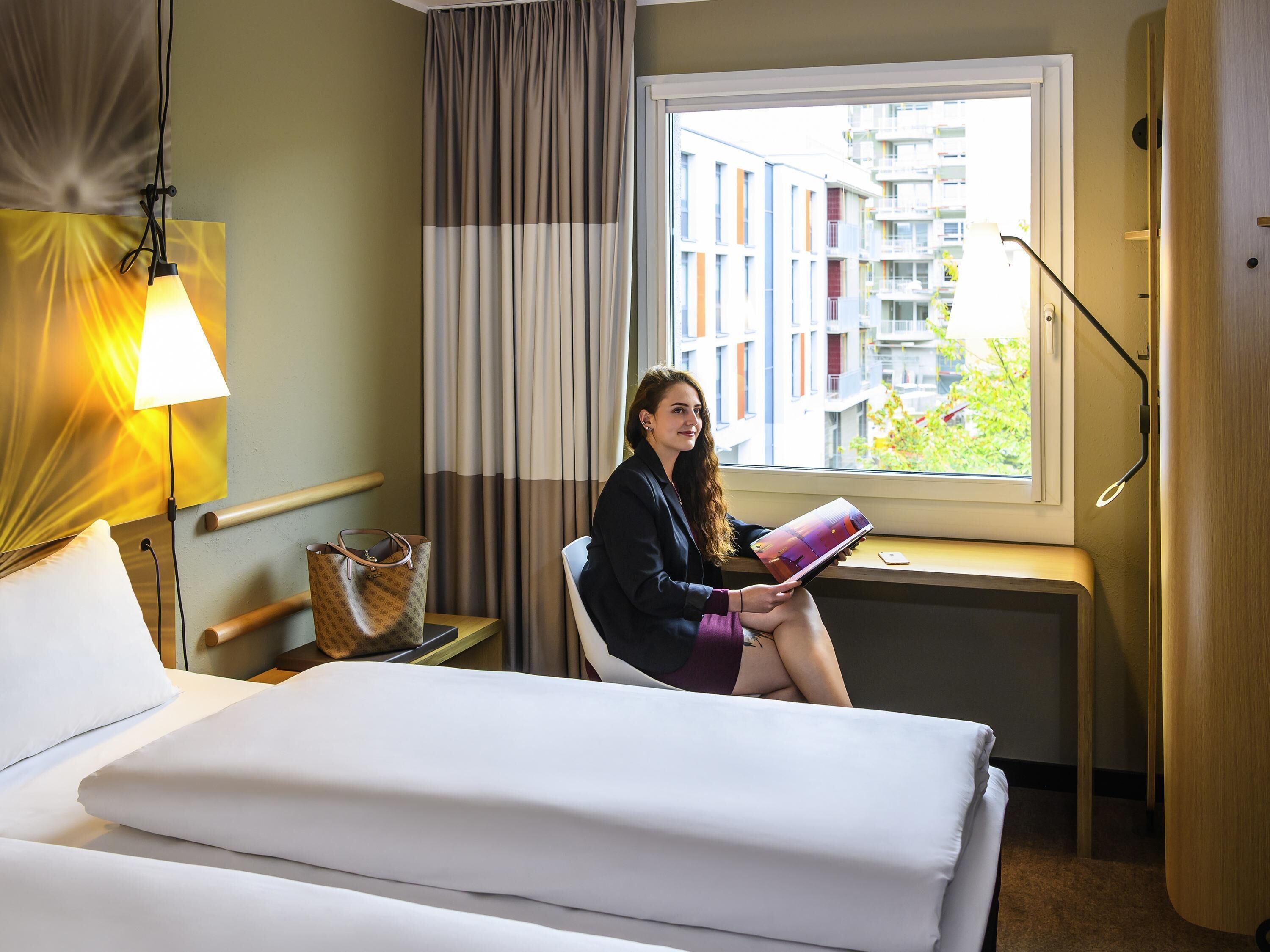 A hotel room with a bed, a desk, a chair, a lamp, and a view of buildings outside the window.