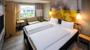 A hotel room with two beds, a desk, a chair, and a window with curtains. - ibis Muenchen City West (Munich)