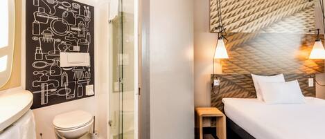 Baño con inodoro, lavamanos y ducha. La habitación tiene una cama con cabecera y lámparas de noche.