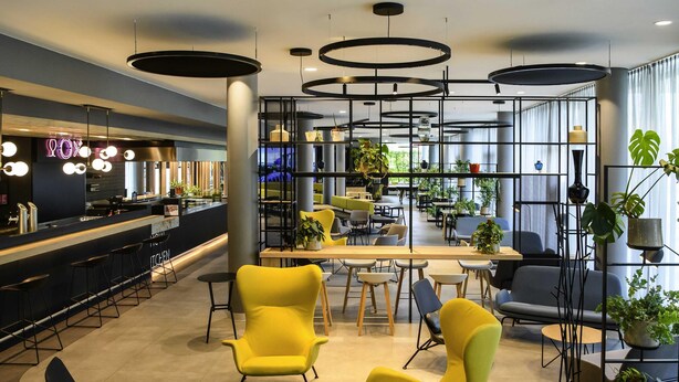 A modern interior with a bar area, yellow chairs, tables, and potted plants. - ibis Muenchen City West (Munich)