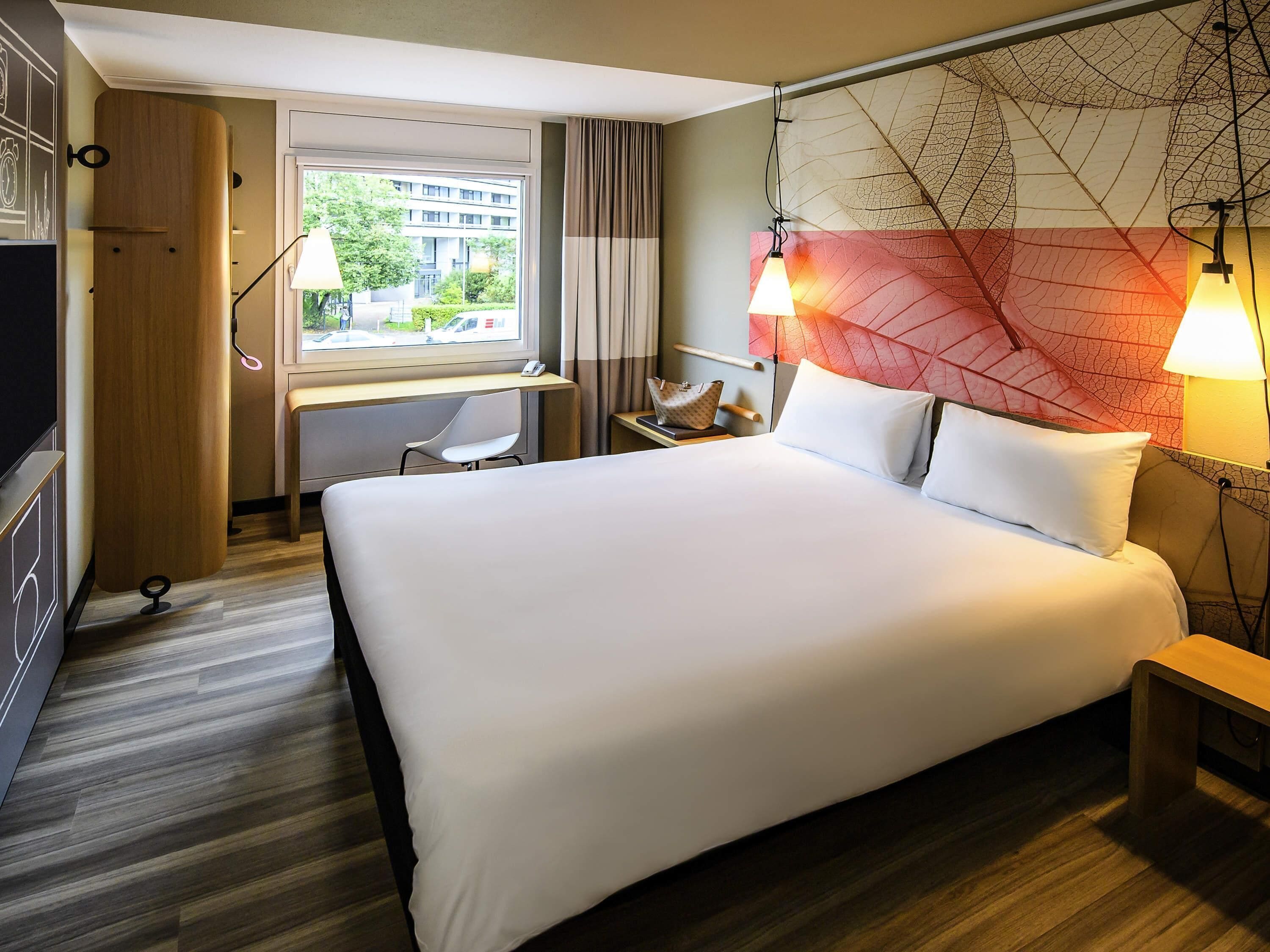 A hotel room with a large bed, a desk, a chair, a window with a view, and two wall-mounted lamps.