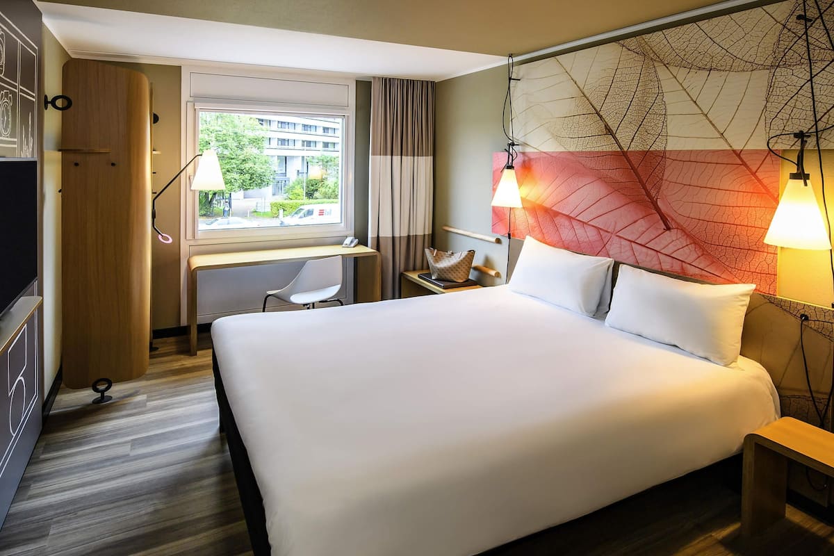 A hotel room with a large bed, a desk, a chair, a window with a view, and two wall-mounted lamps.