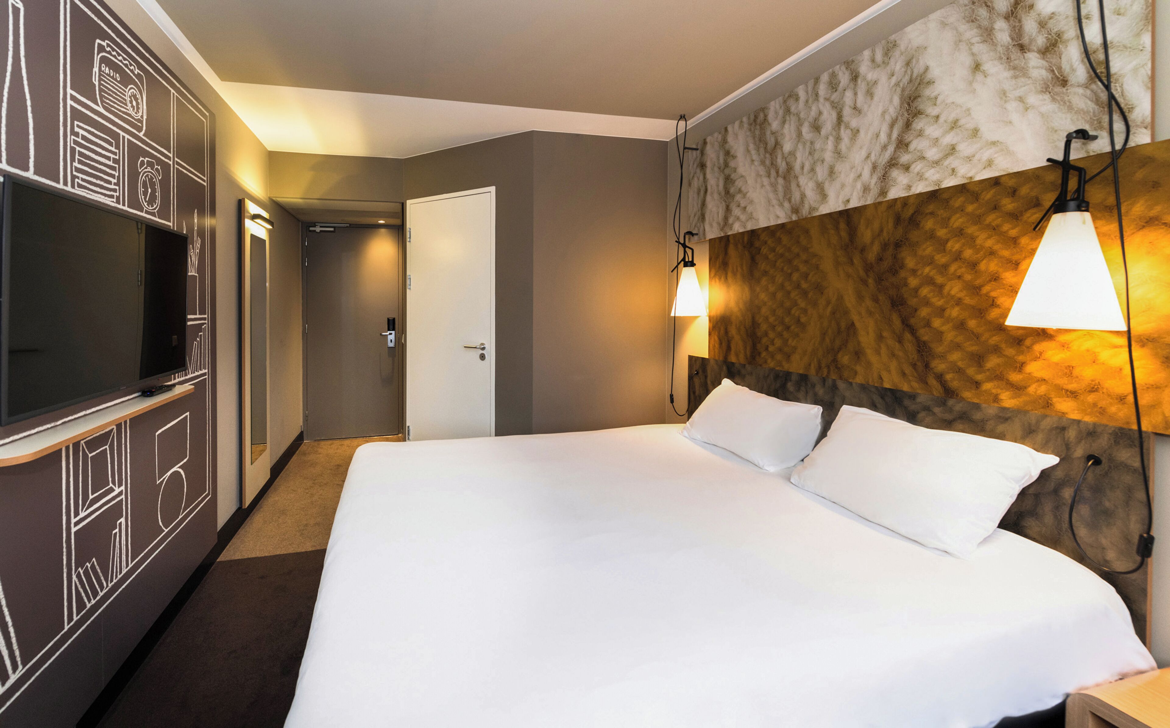 A hotel room with a large bed, a television on a stand, a door to a hallway, and a wall-mounted lamp.