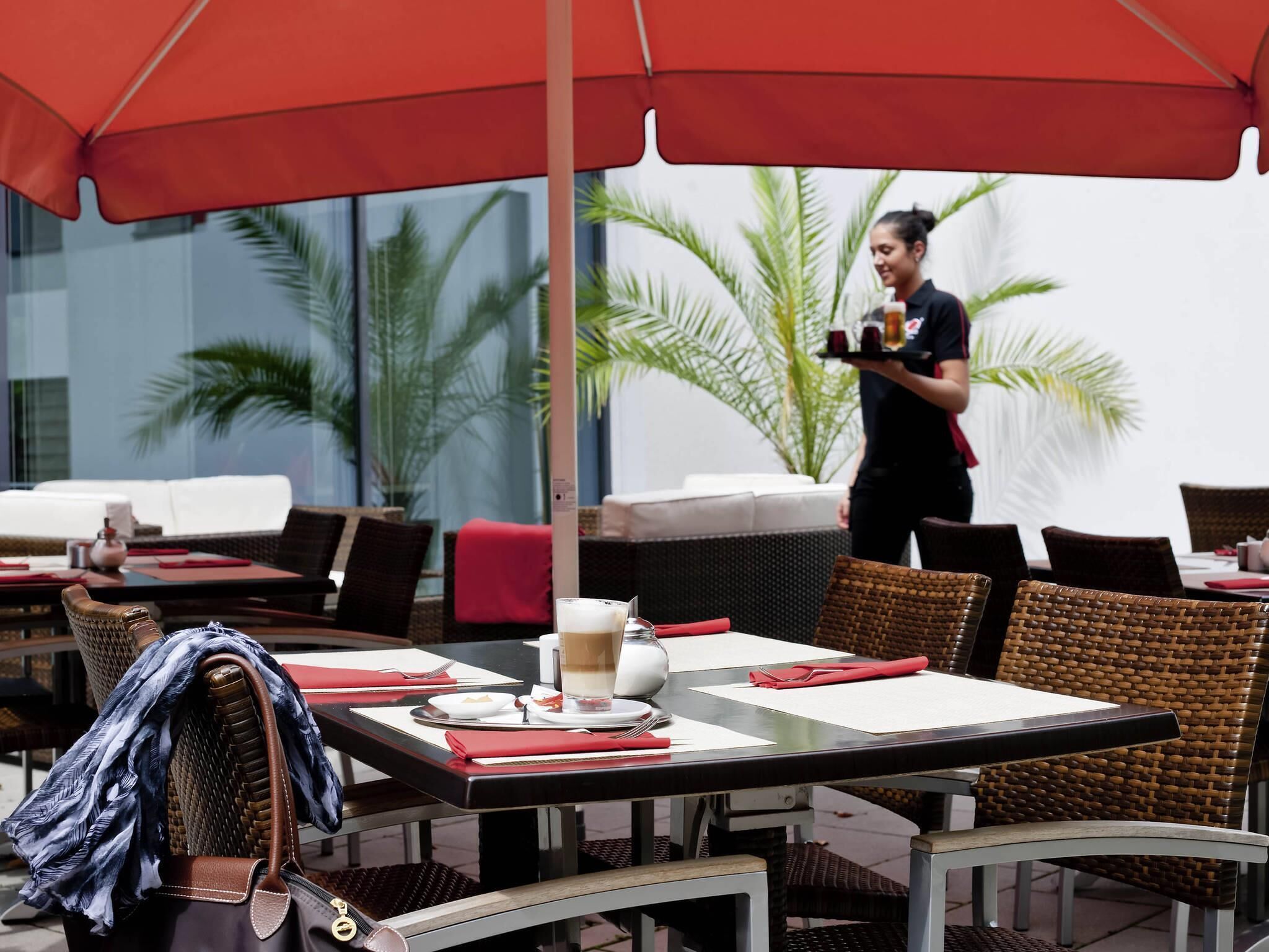 An outdoor dining area with wicker furniture, red umbrellas, and a waiter serving.
