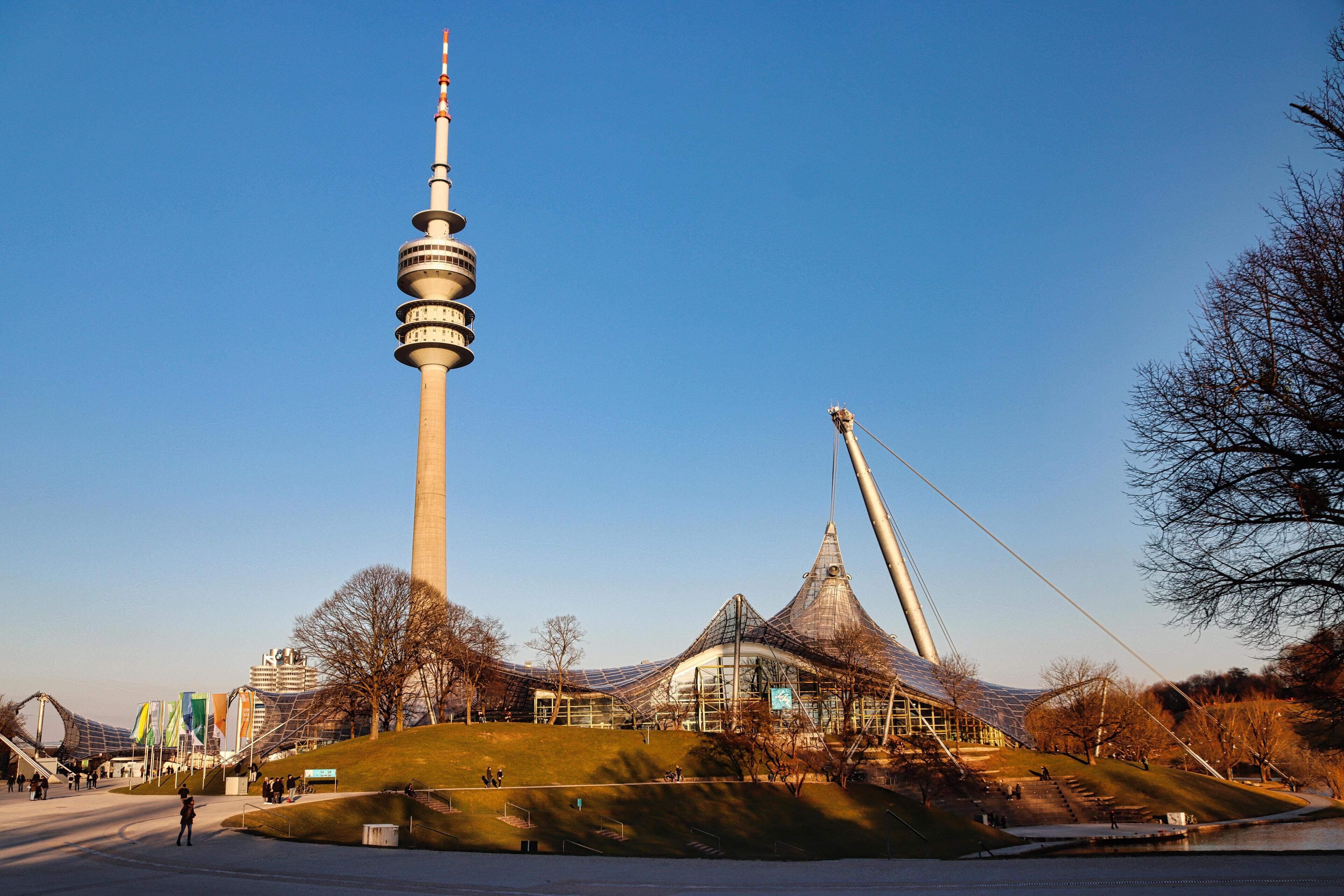 A large tower and a distinctive building with a curved roof, surrounded by a park with a small pond and pathways.
