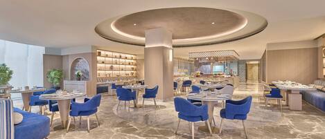 A spacious hotel lobby with multiple seating areas, a central column, and a view into the dining area.