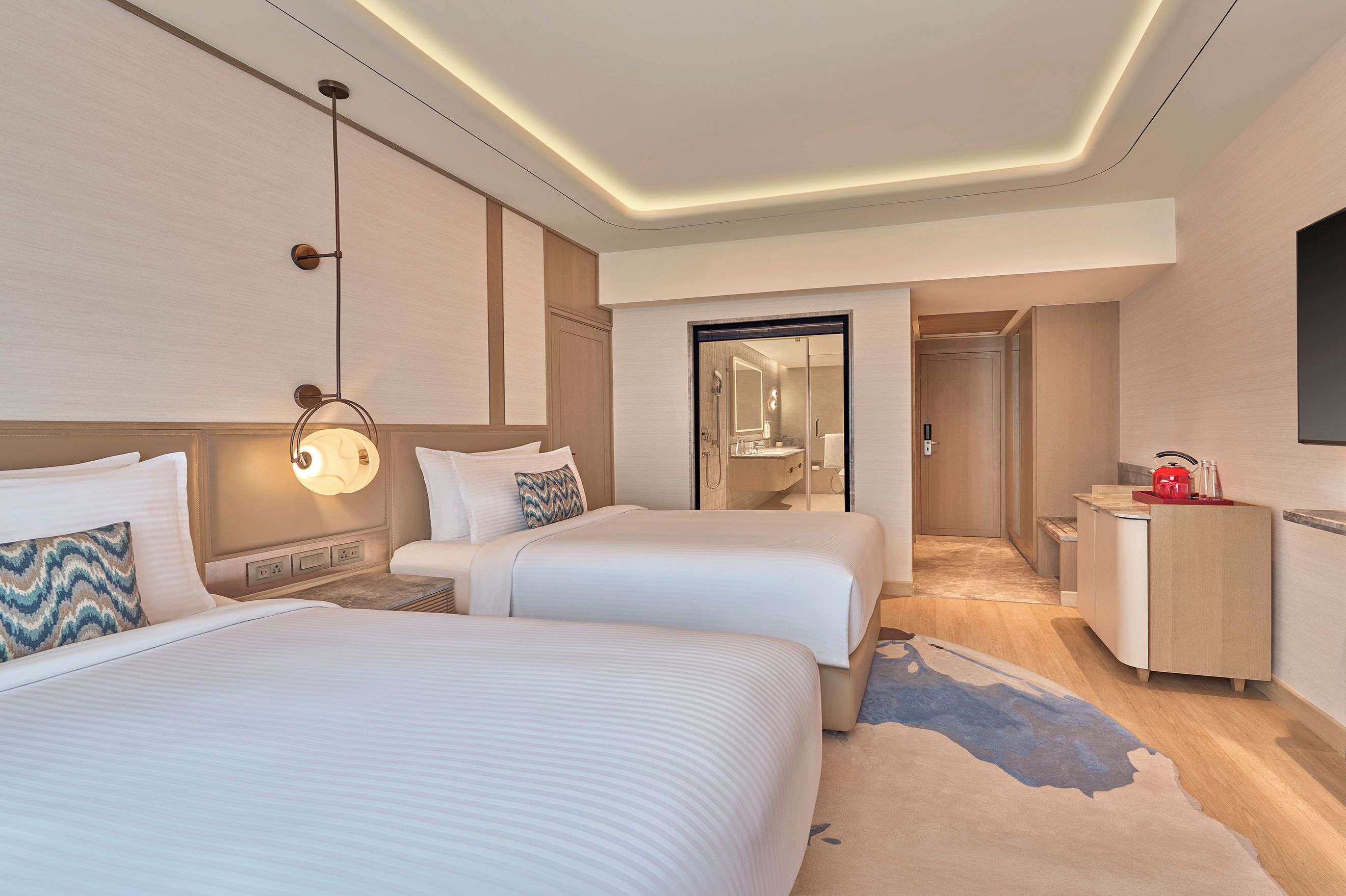 Habitación de hotel con dos camas, un escritorio, un espejo y un baño visible a través de una puerta abierta.