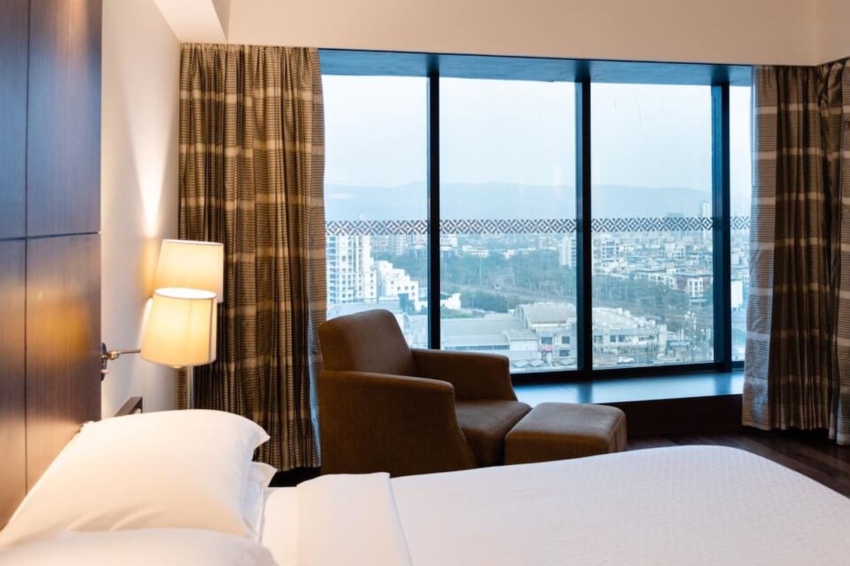 Habitación de hotel con una cama grande, una silla, un escritorio y vistas a un paisaje urbano a través de amplios ventanales.