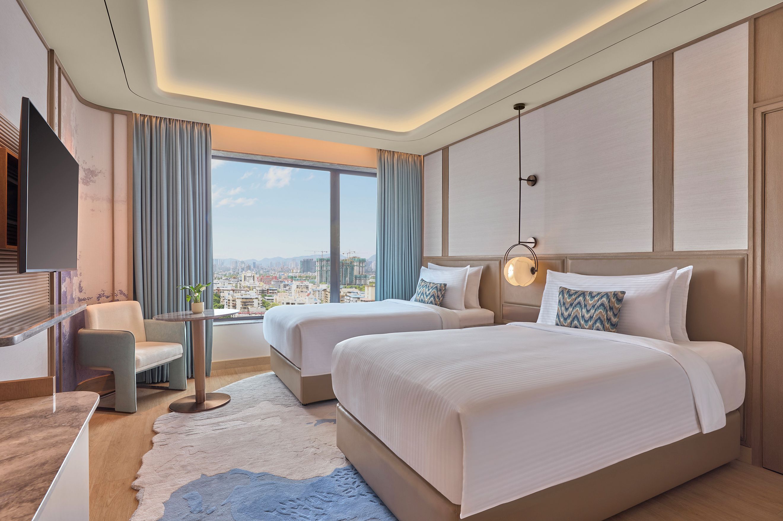 Habitación de hotel con dos camas, un escritorio, una silla, un televisor y un ventanal con vistas a la ciudad.