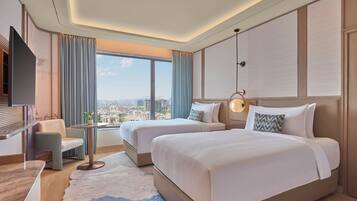 Habitación de hotel con dos camas, un escritorio, una silla, un televisor y un ventanal con vistas a la ciudad.