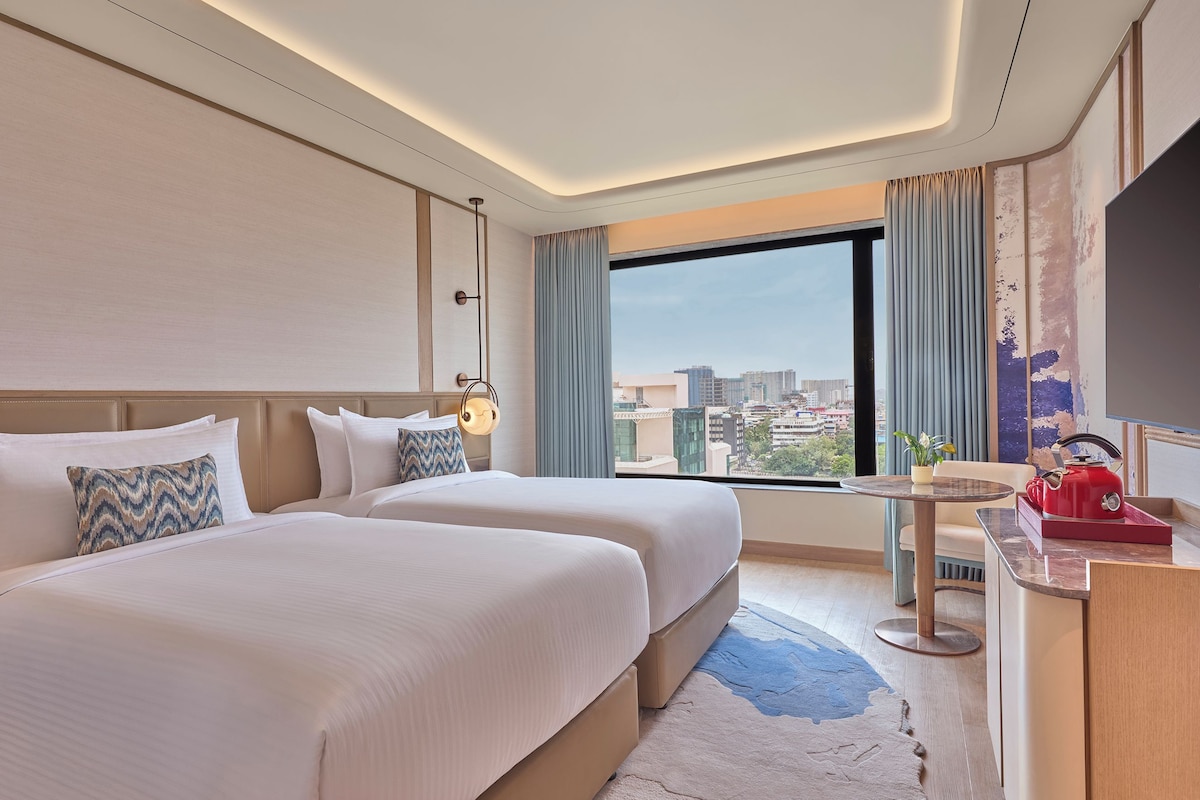 Habitación de hotel con dos camas, un escritorio con hervidor rojo, vista a la ciudad y un amplio ventanal con cortinas.