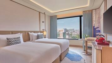 Ein Hotelzimmer mit zwei Betten, einem Schreibtisch mit einem roten Wasserkocher und einem großen Fenster mit Blick auf die Stadt.