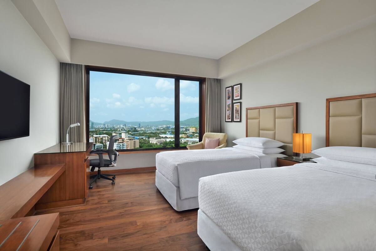 A hotel room with two beds, a desk, a chair, a TV, and a view of a cityscape.