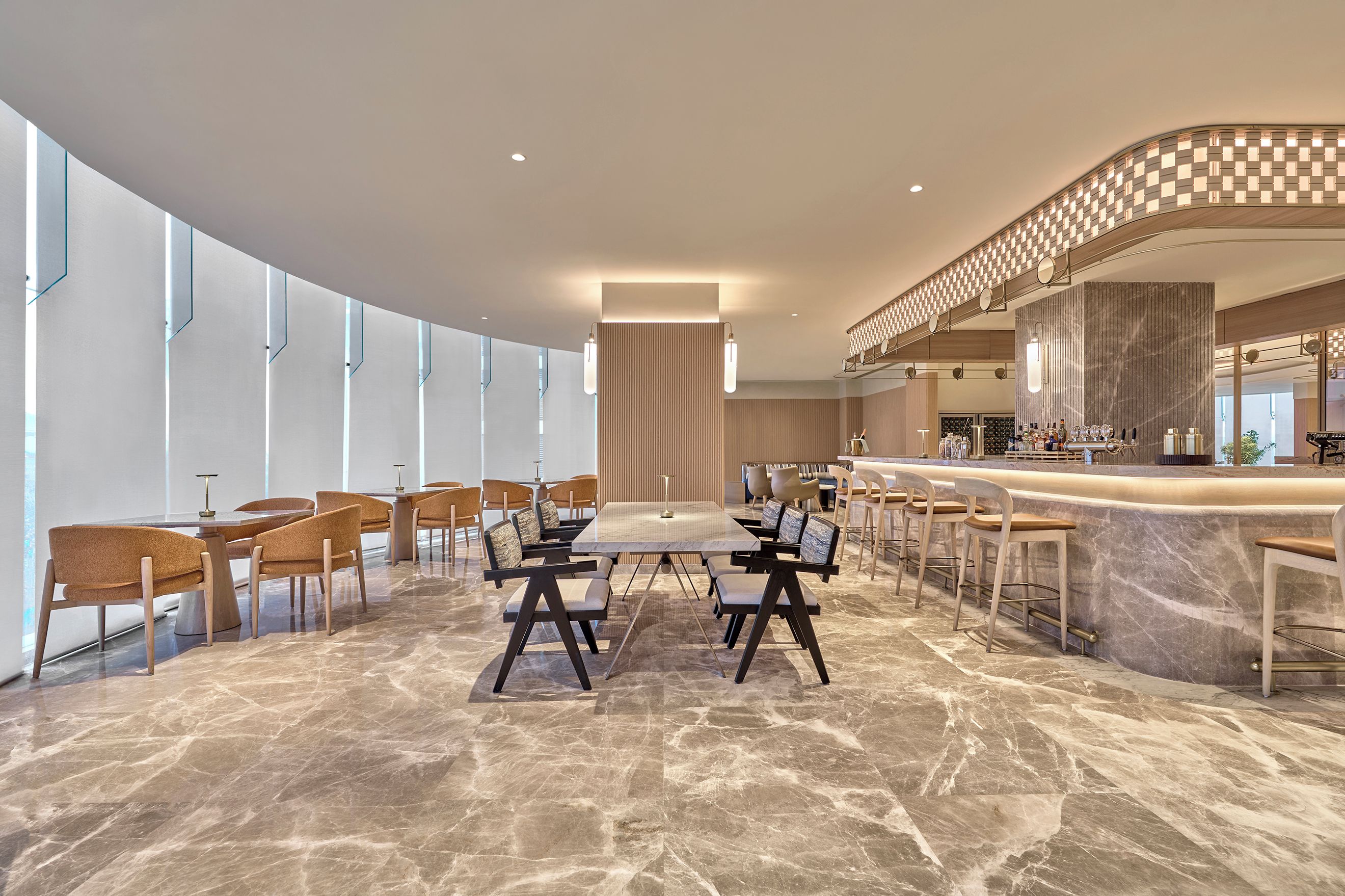 Un moderno vestíbulo de hotel con piso de mármol, un área de comedor central con mesas y sillas, un bar con taburetes y un ventanal que proporciona luz natural.