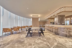 A modern hotel lobby with a marble floor, a central dining area with tables and chairs, a bar area with stools, and a large window providing natural light.