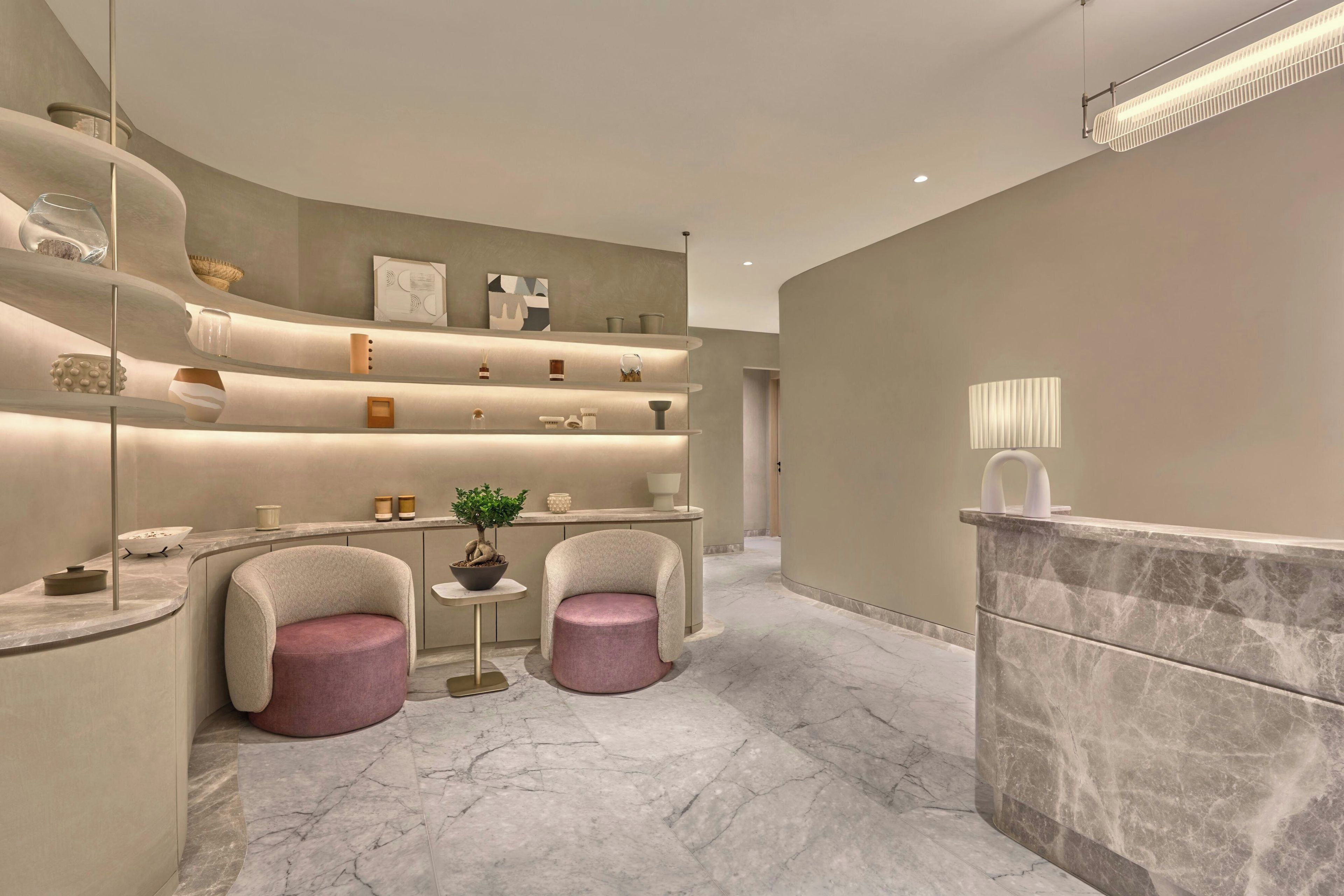 Un interior moderno con un mostrador de recepción, dos sillones, una pequeña mesa con una planta y estantes con diversos objetos.