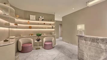 Un interior moderno con un mostrador de recepción, dos sillones, una pequeña mesa con una planta y estantes con diversos objetos.