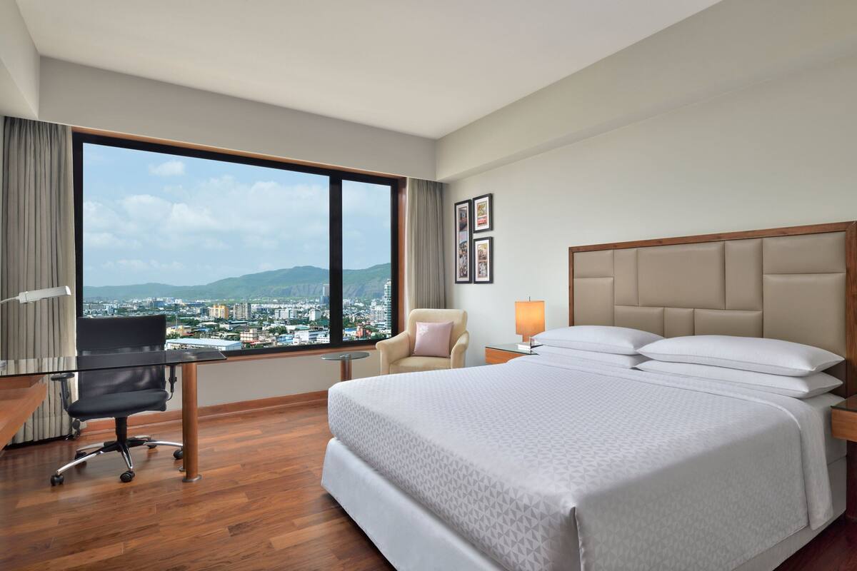 A hotel room with a large bed, a desk with a chair, a lamp, and a view of a cityscape through a large window.