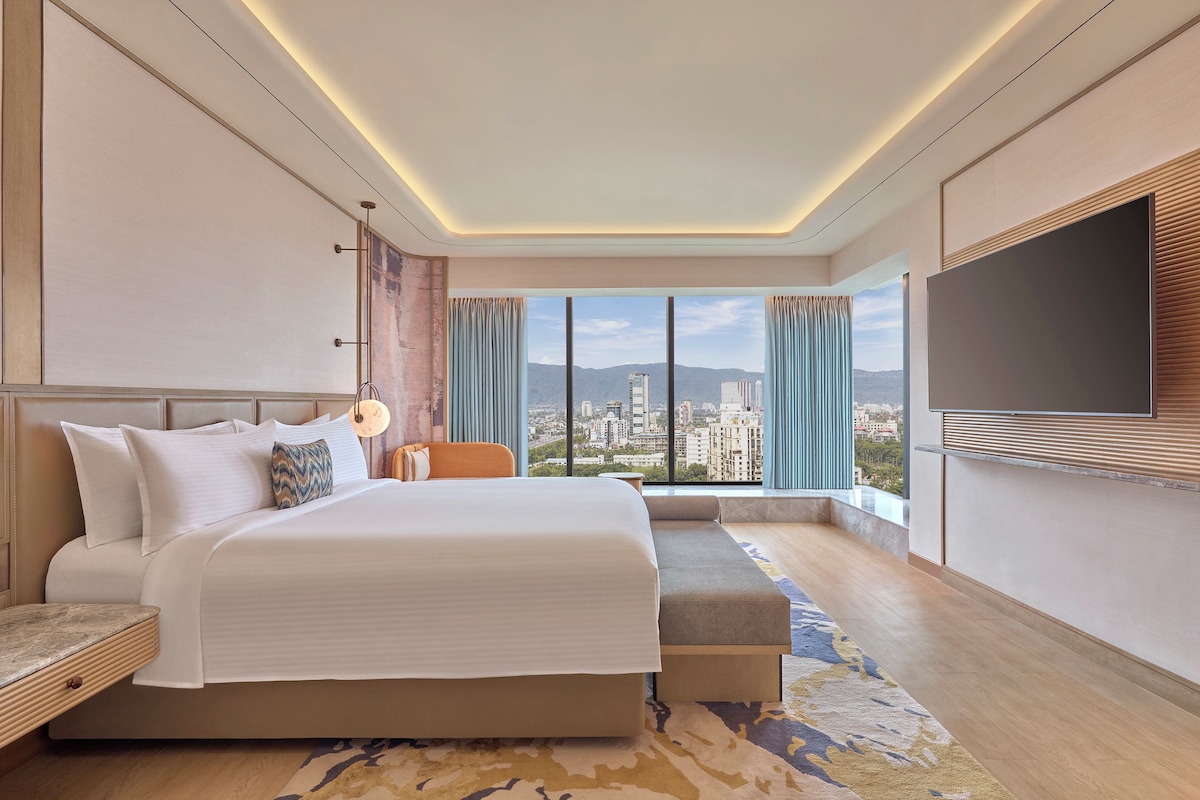 Una habitación de hotel moderna con una cama grande, zona de estar, televisor y vista a la ciudad.