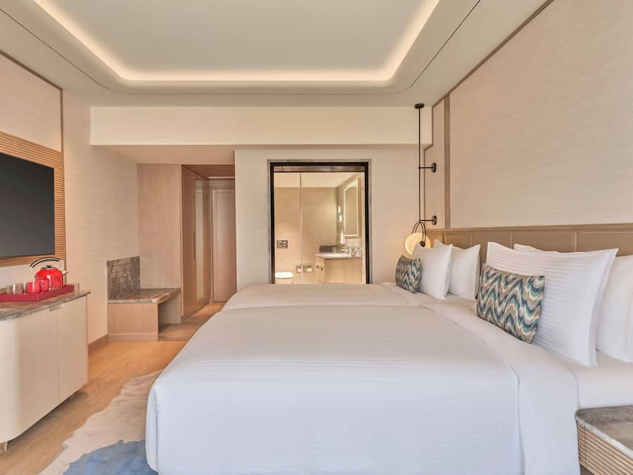 Habitación de hotel con una cama grande, televisión, escritorio con hervidor y baño visible a través de una puerta abierta.