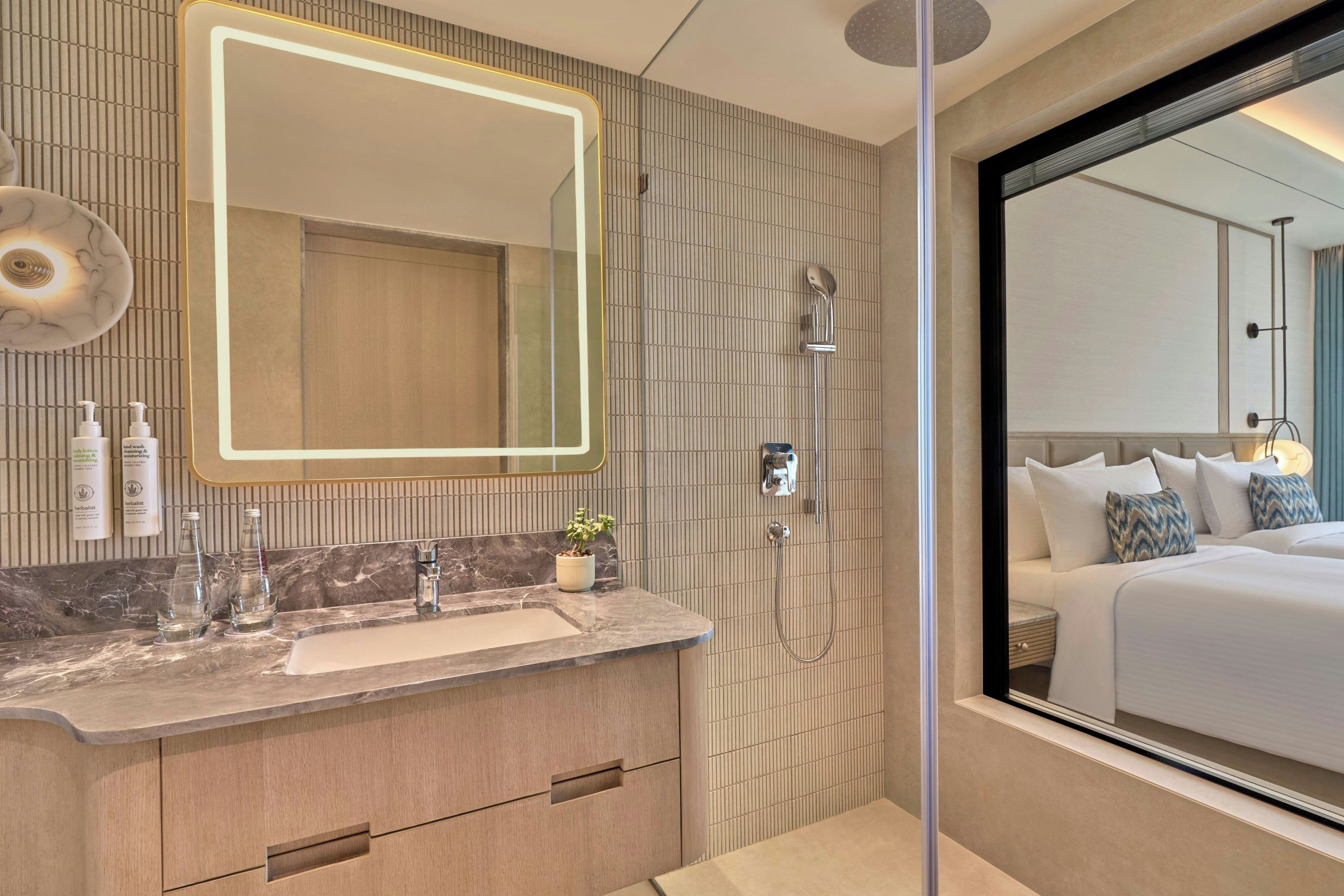 Un baño moderno con tocador, un espejo grande, una ducha y vistas a un dormitorio con cama y almohadas.