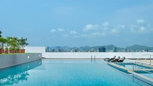 Ein Pool auf dem Dach mit klaren blauen Fliesen, einer weißen Wand und Blick auf die Stadt im Hintergrund.