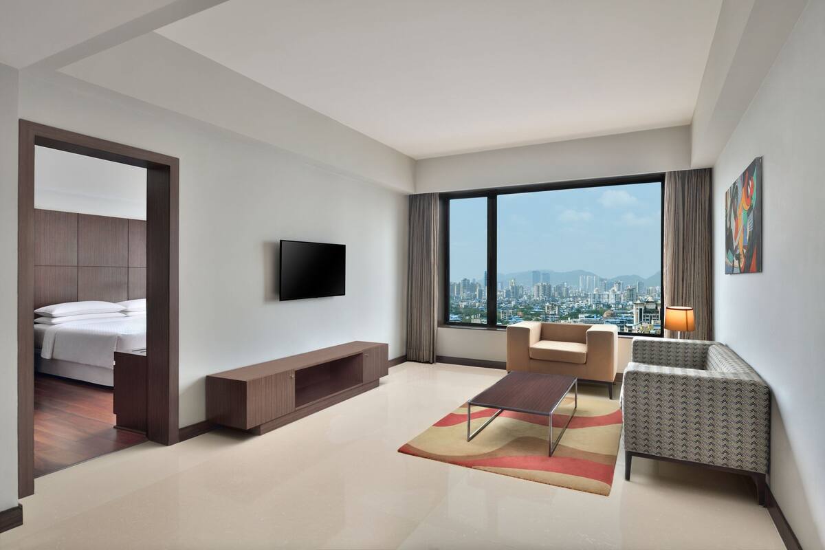A modern hotel room with a large window, a bed, a TV, a sofa, and a coffee table.