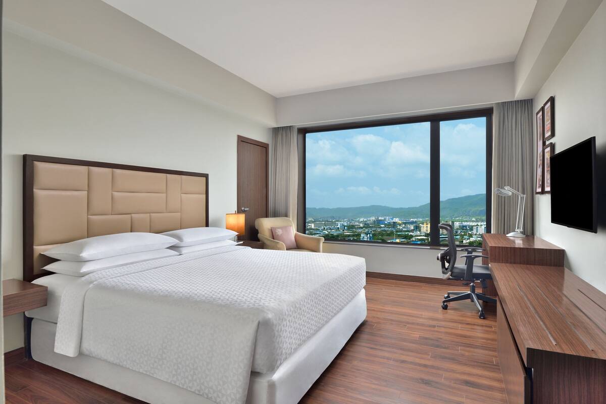 A hotel room with a large bed, a desk, a chair, a TV, and a window with a city view.