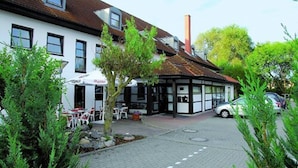 Een gebouw met een witte gevel, meerdere ramen en een pannendak. Er is een klein terras met tafels en stoelen.