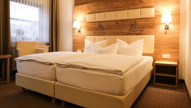 Een tweepersoonsbed met houten hoofdeinde, twee nachtkastjes, een stoel en een raam met dunne gordijnen.